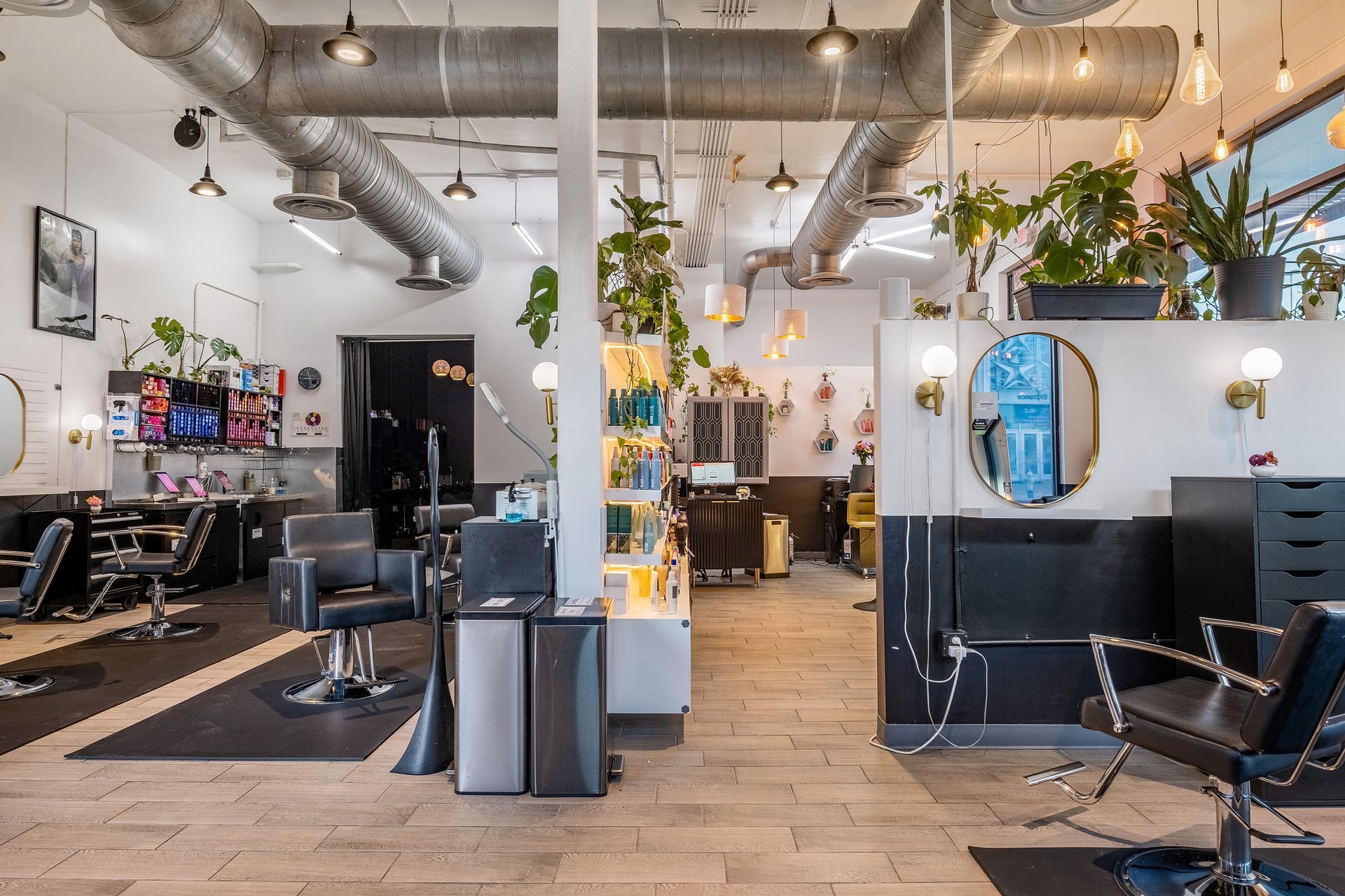 Interior of a hair salon with chairs, mirrors, plants, and product displays. White walls, black accents, and exposed ductwork.