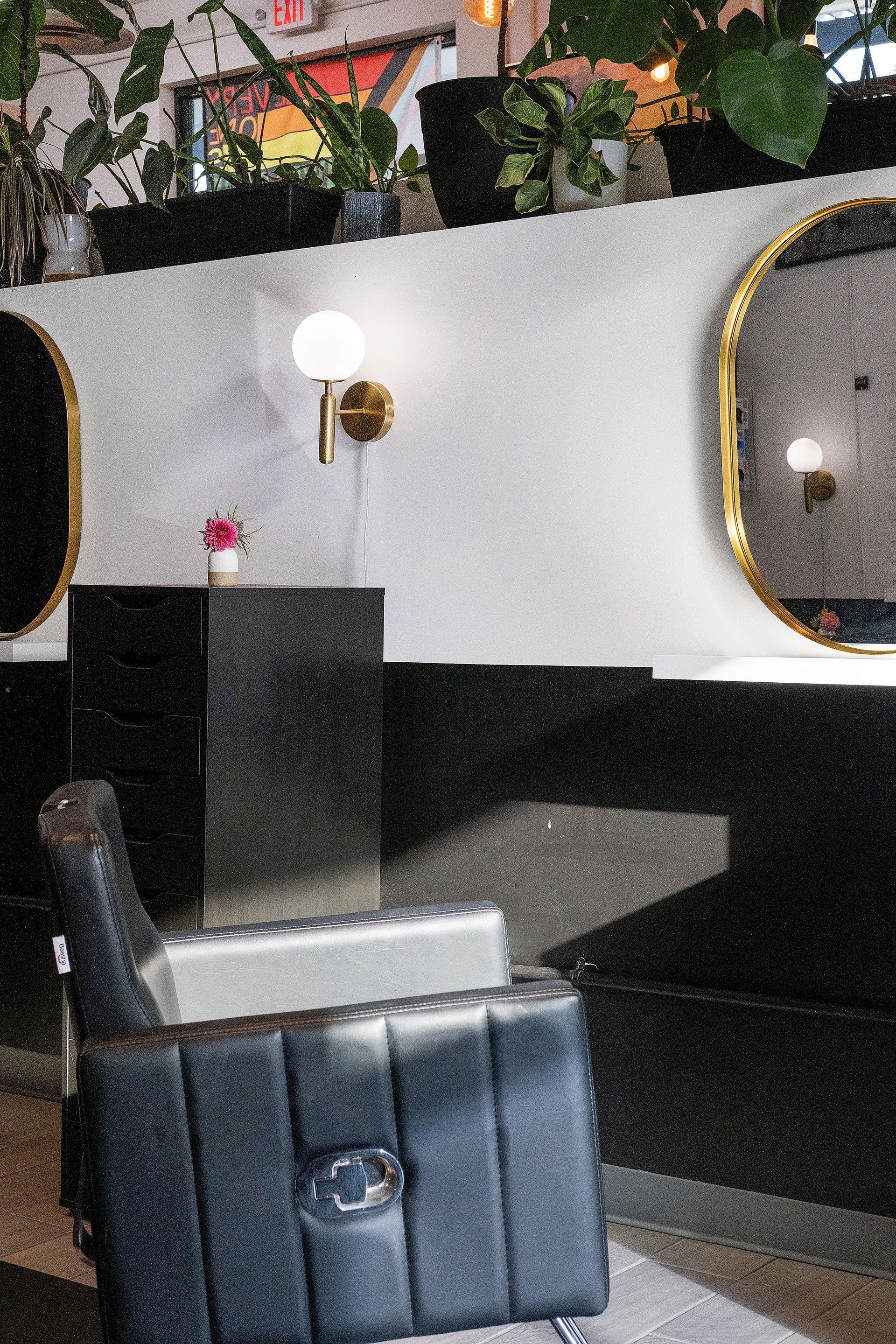 A black leather barber chair, a black pedestal, and a gold-framed mirror are in a salon with plants.