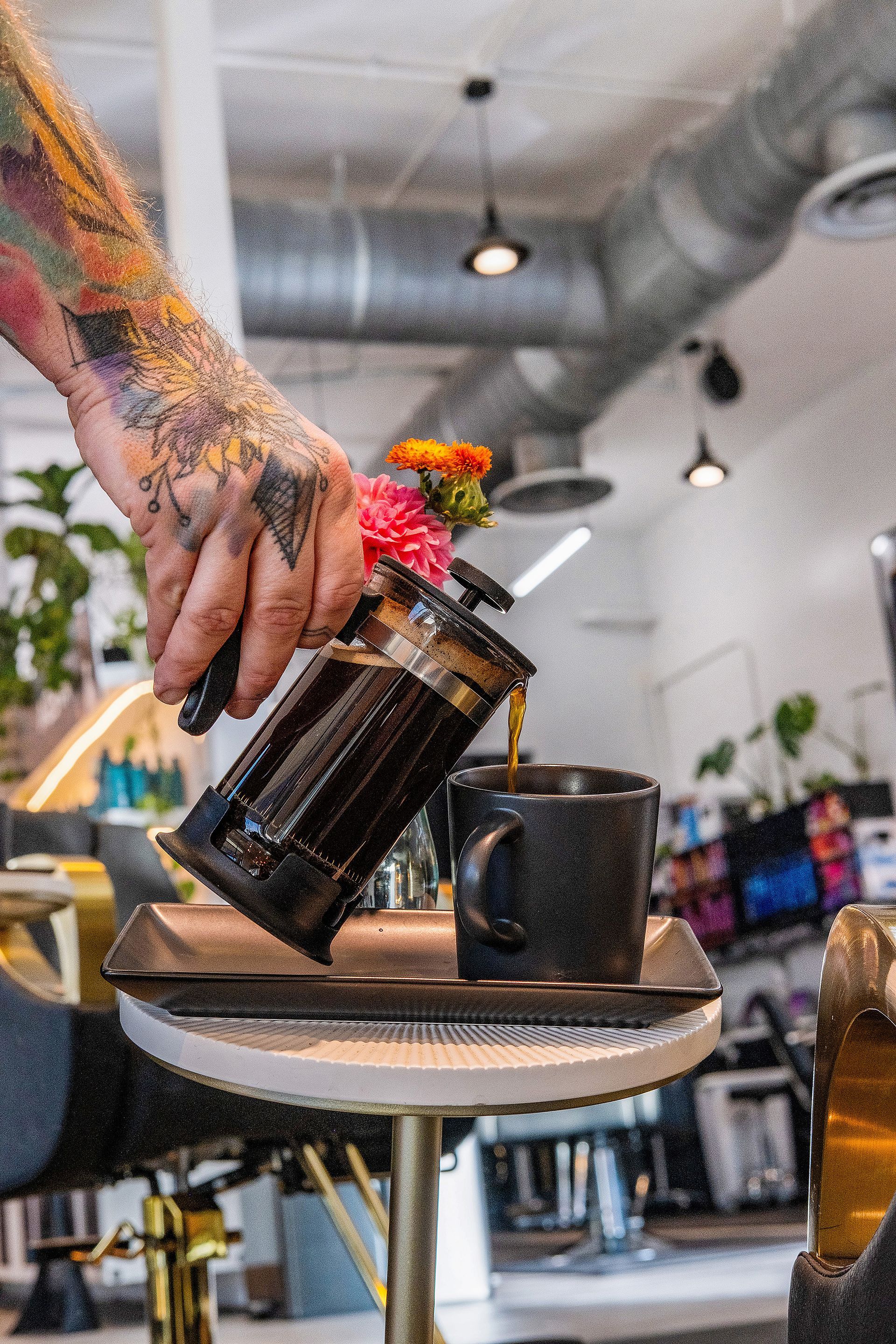 Person pouring coffee from a French press into a mug, indoors, with tattoos visible.