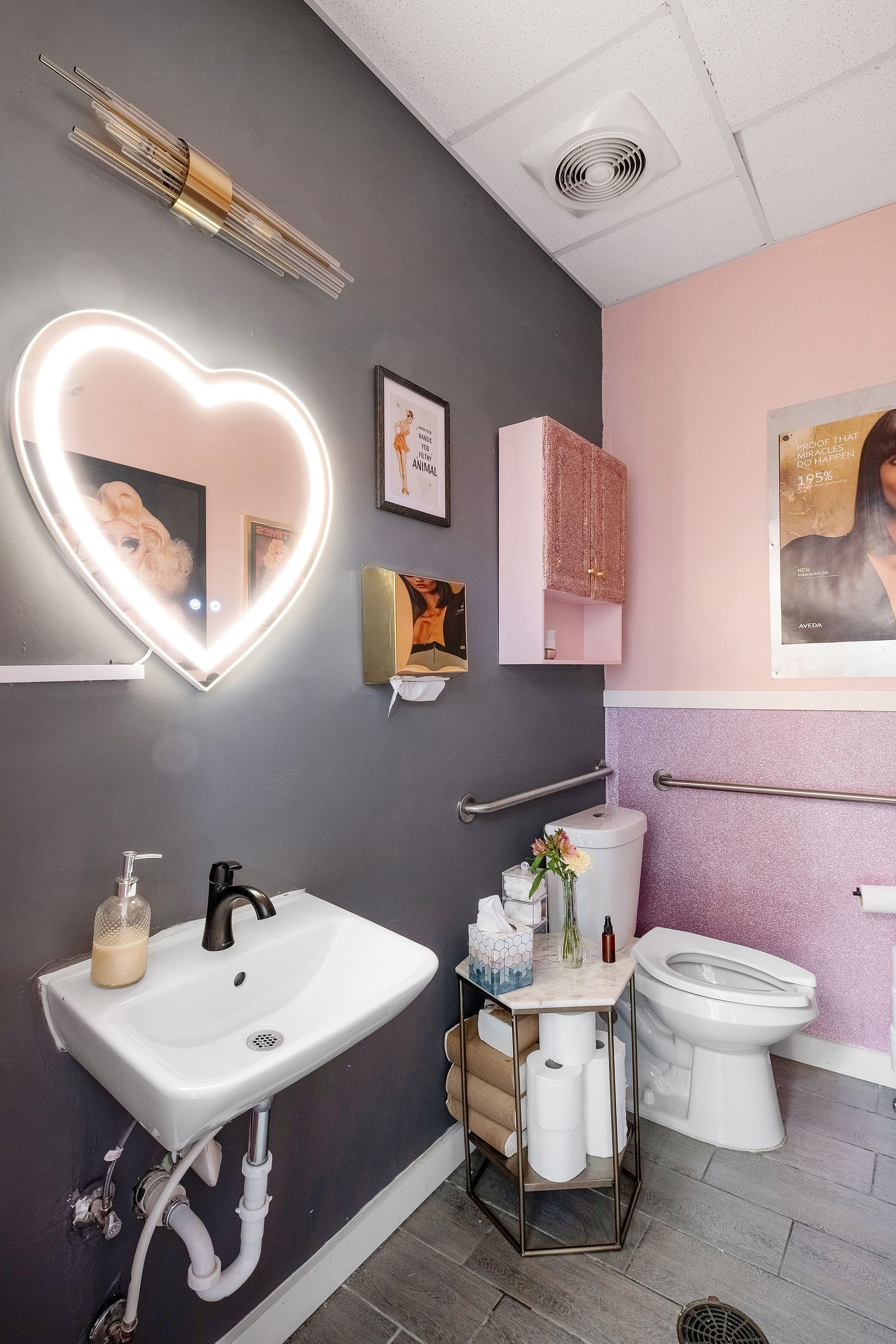 A small bathroom with a heart-shaped mirror, pink and gray walls, and a white sink and toilet.