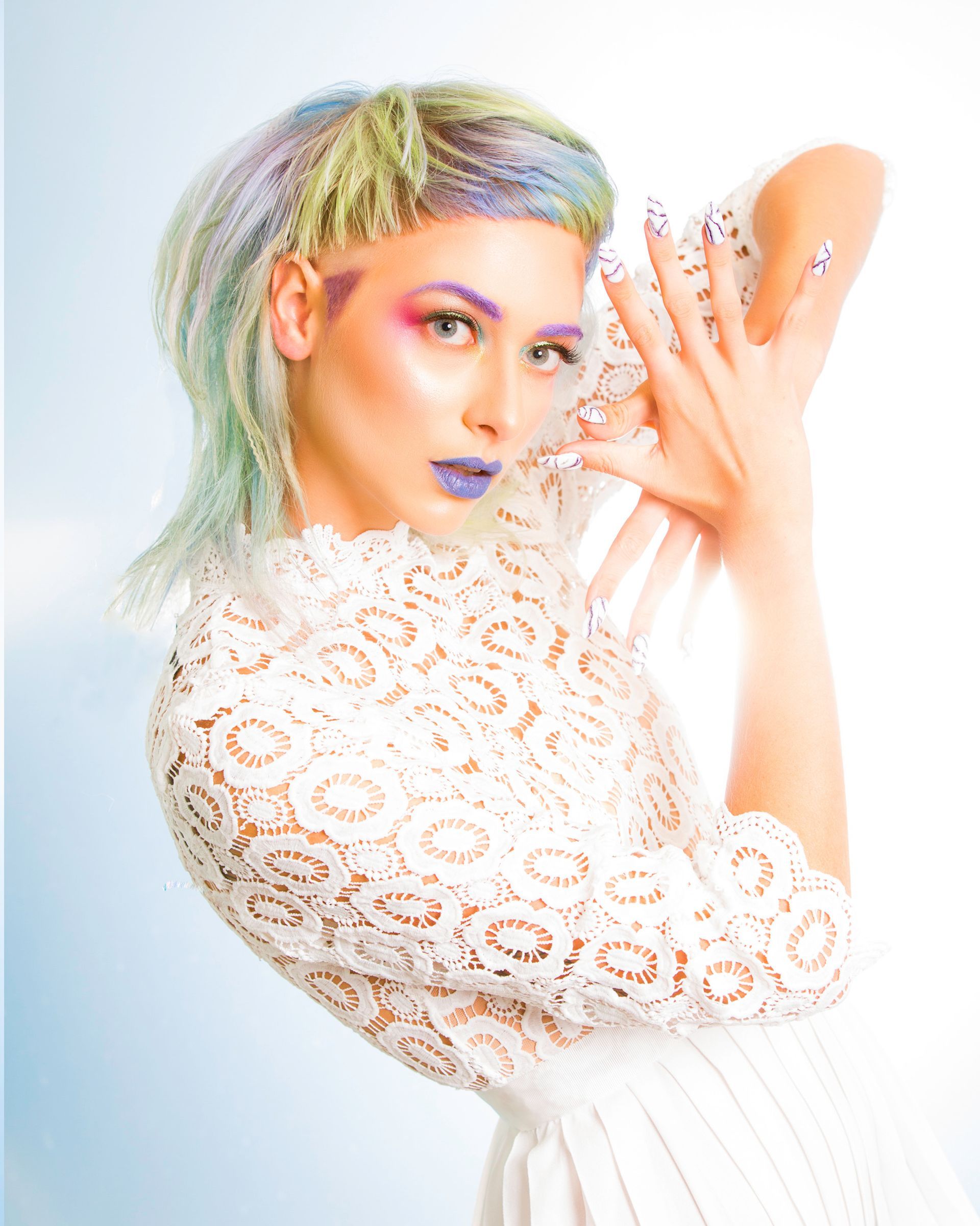 A woman with colorful hair and nails is wearing a white lace top.