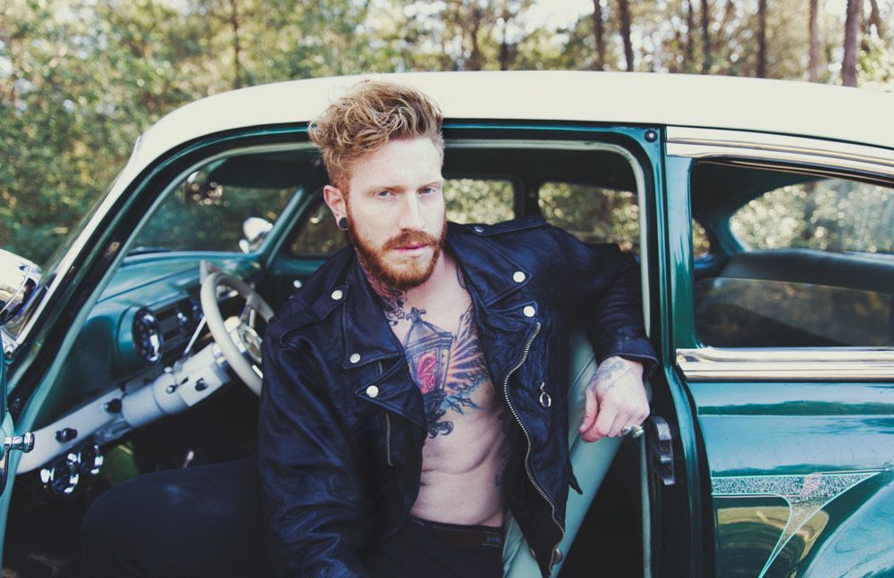 A shirtless man with a tattoo on his chest is sitting in a green car.