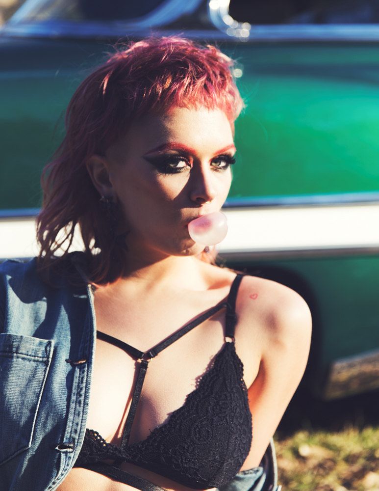 A woman with pink hair blowing a bubble in front of a green car