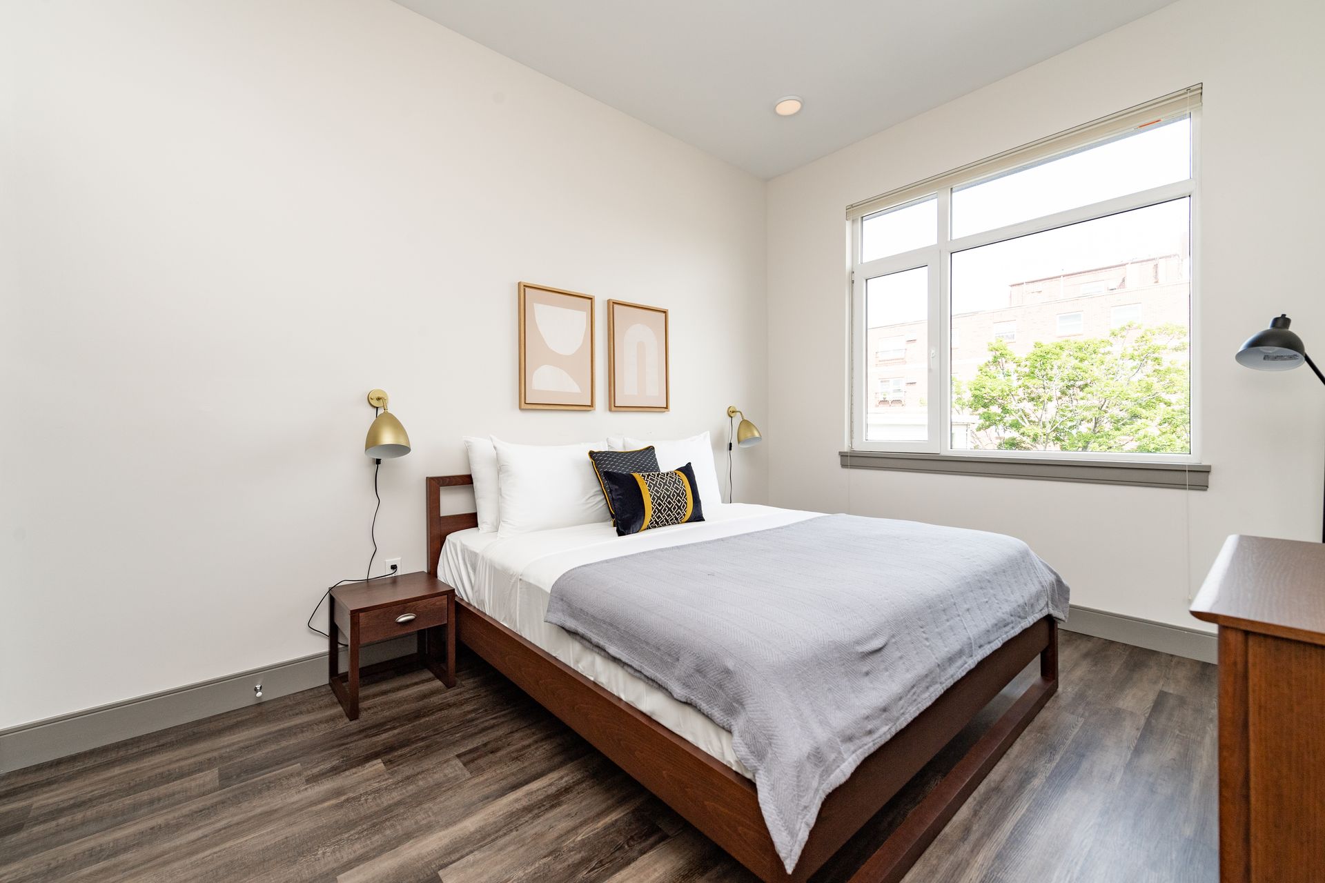 Bedroom with a bed, nightstand, dresser, and window. Gray flooring, white walls, and wood furniture.