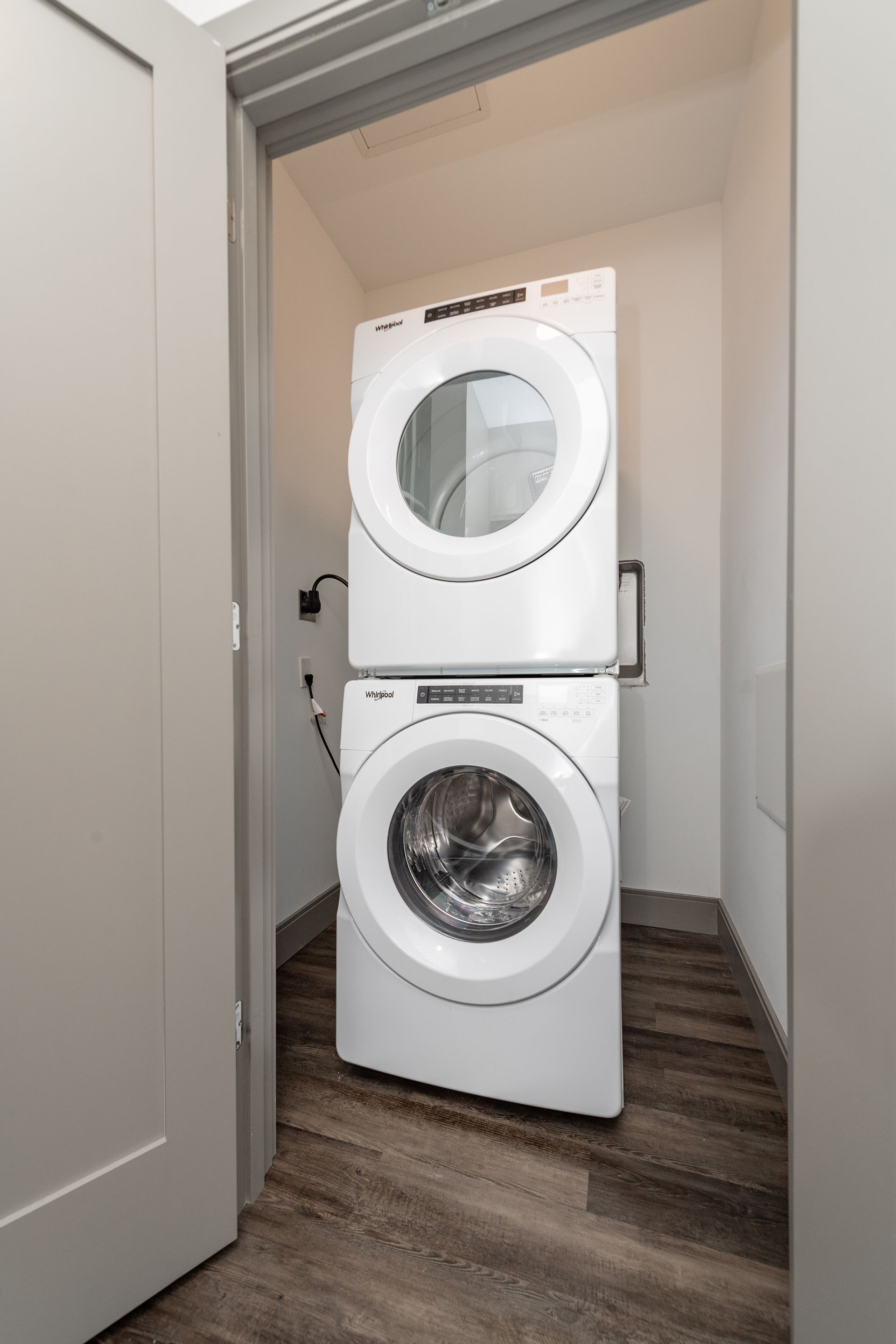 Stacked white washer and dryer in a small laundry room with wood-look flooring.