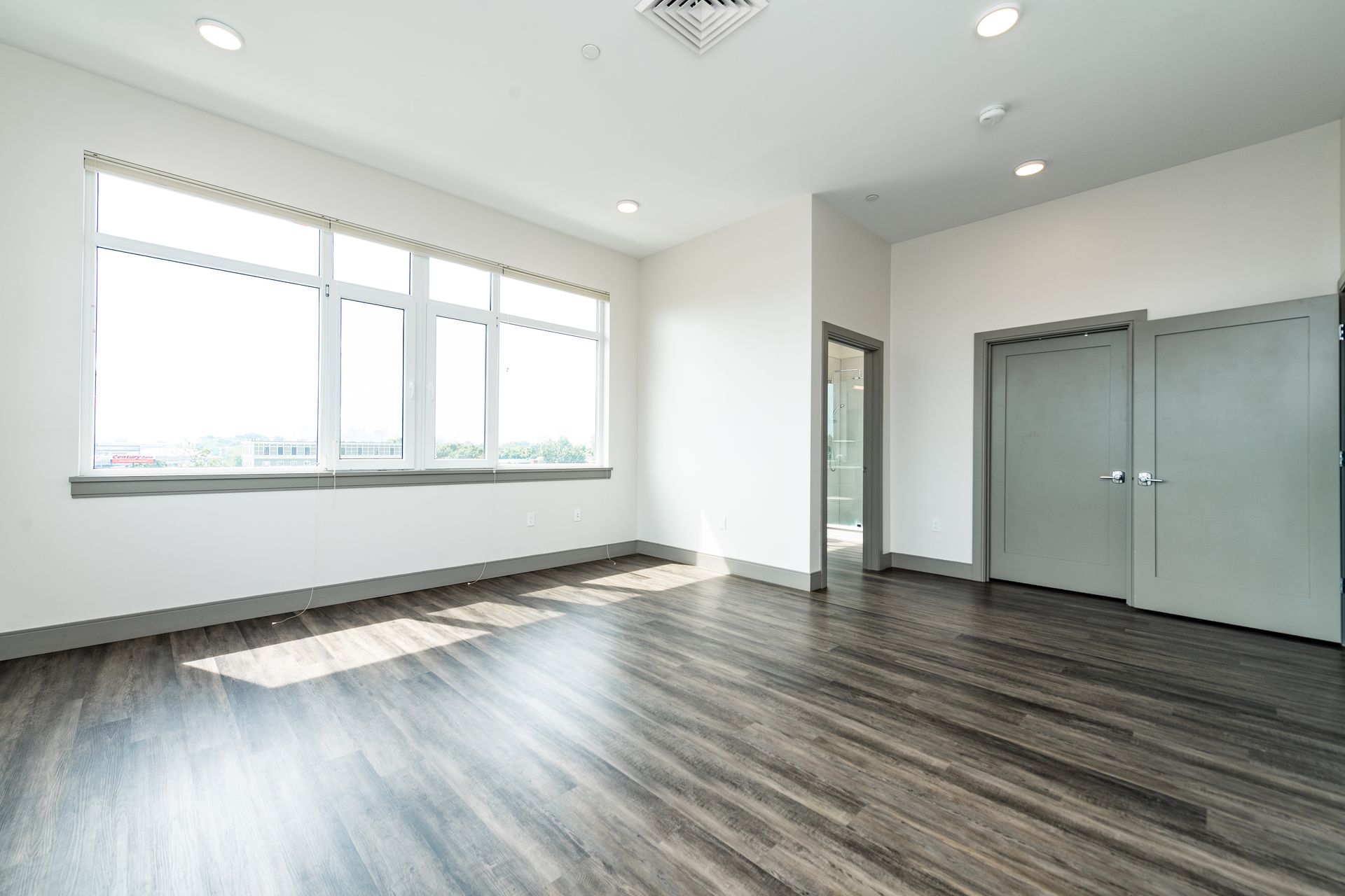 Empty, modern room with large windows, dark wood-look flooring, and gray doors.