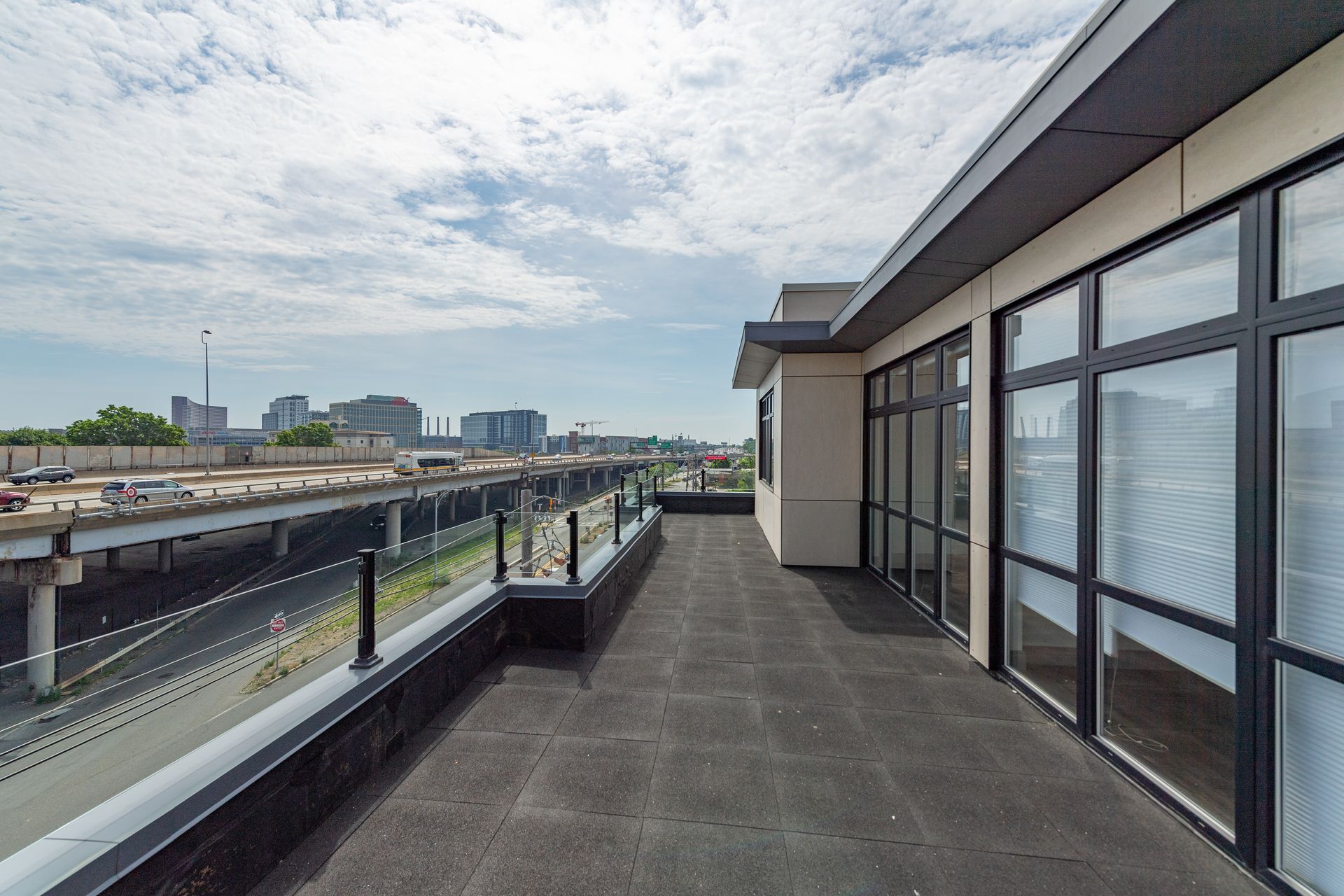 Rooftop patio with glass railing, overlooking a highway and city skyline under a cloudy sky.