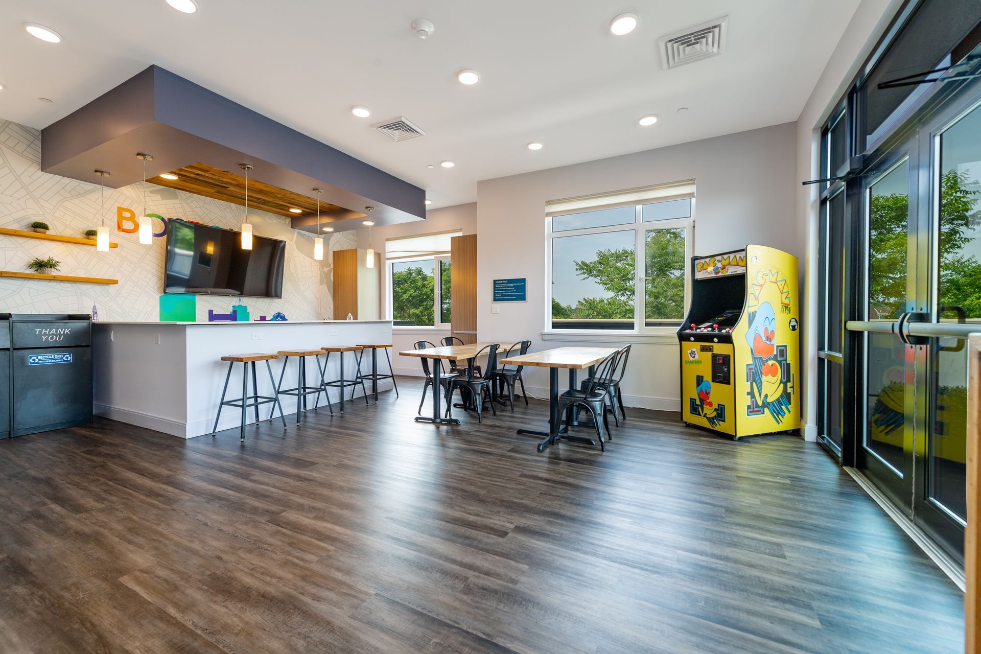 Recreational room with bar seating, tables, arcade game, windows overlooking greenery, and a dark wood floor.
