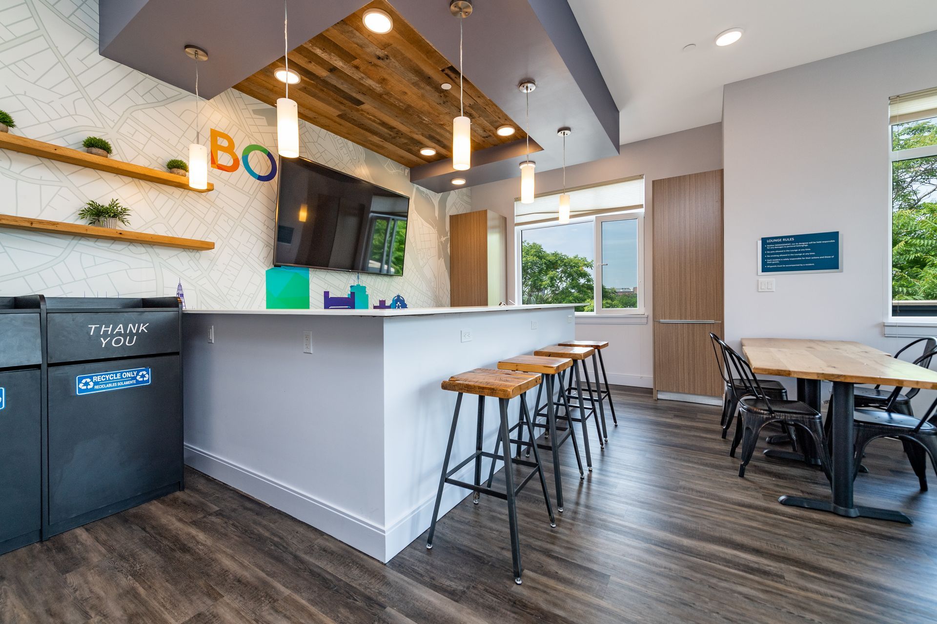 Communal lounge with bar seating, table, TV, shelves, and trash/recycling bins. Neutral colors, natural light.