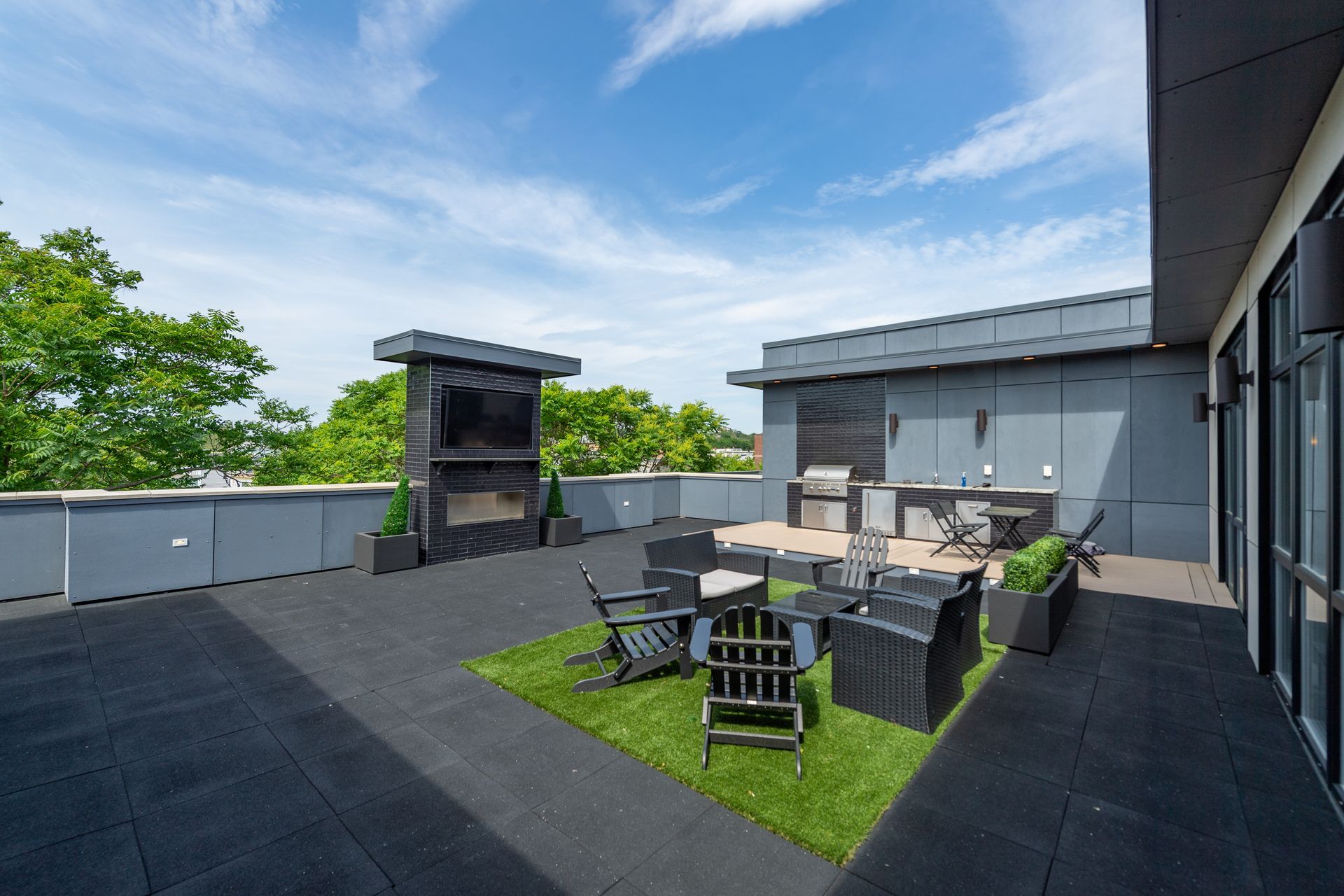 Rooftop patio with outdoor kitchen, TV, and seating area. Overcast sky, trees in the background.