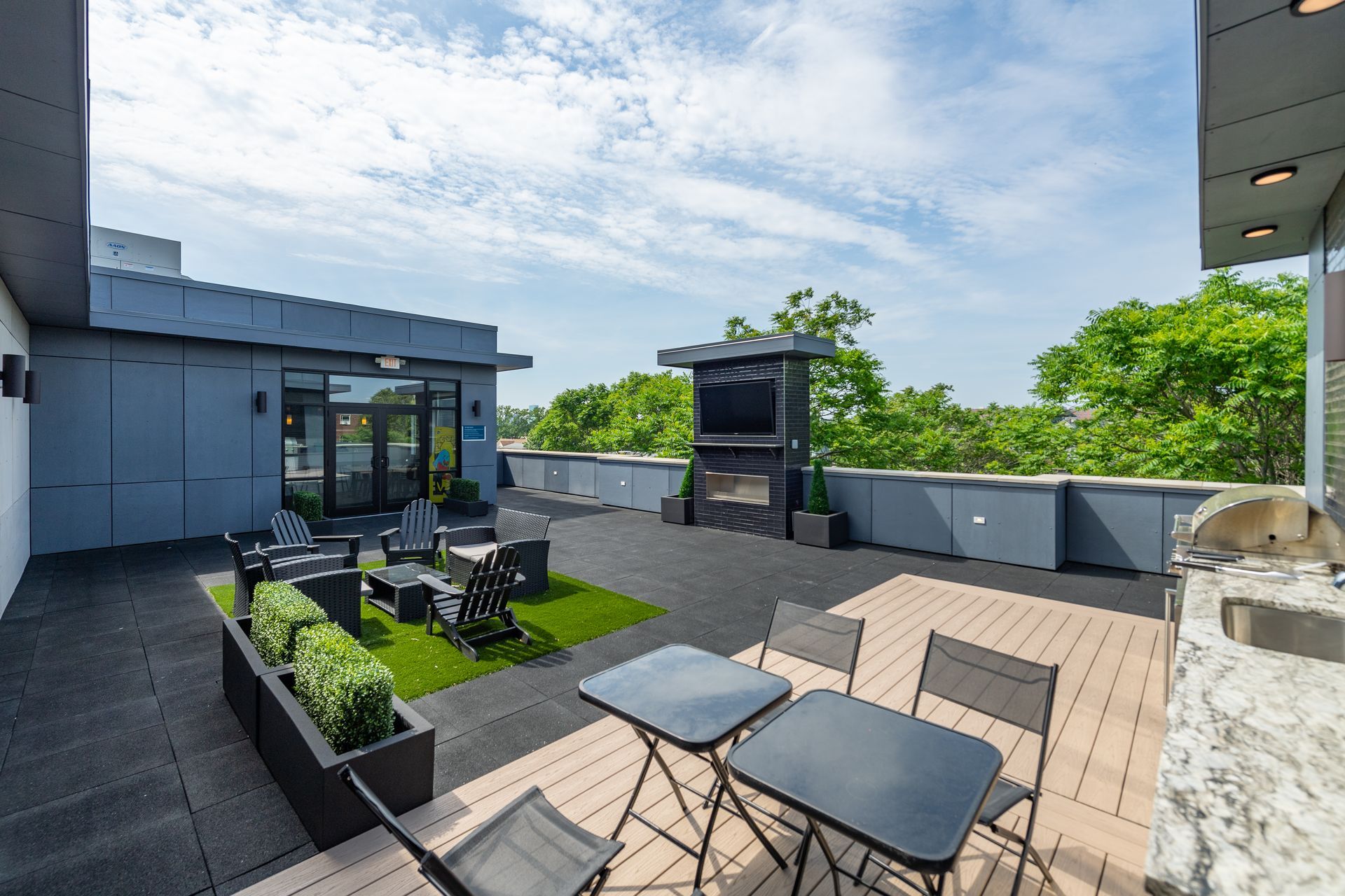 Rooftop deck with seating, fireplace, and greenery. Blue sky, neutral tones.