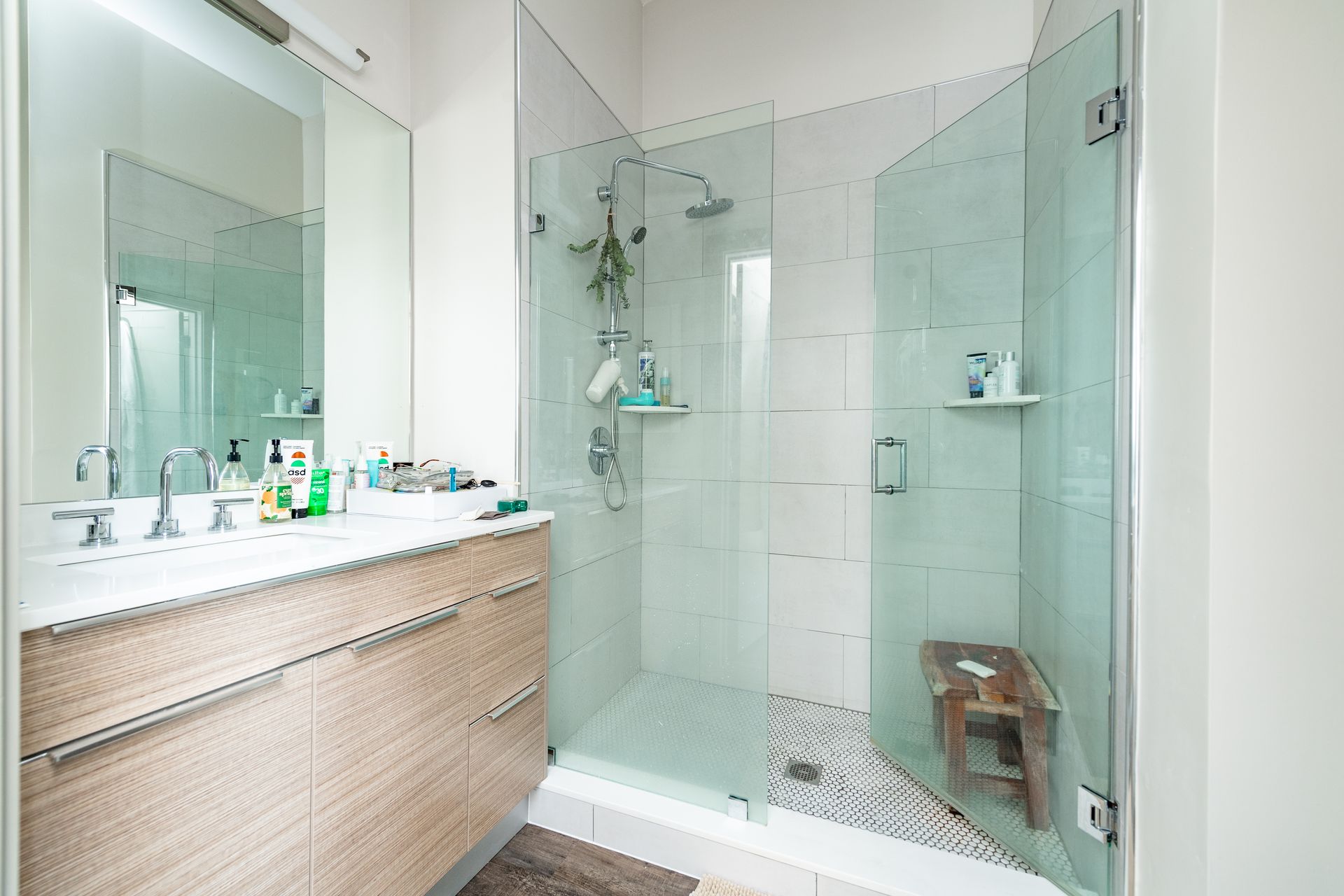 Bathroom with glass-enclosed shower, white tile, vanity, mirror, and a small wooden stool.