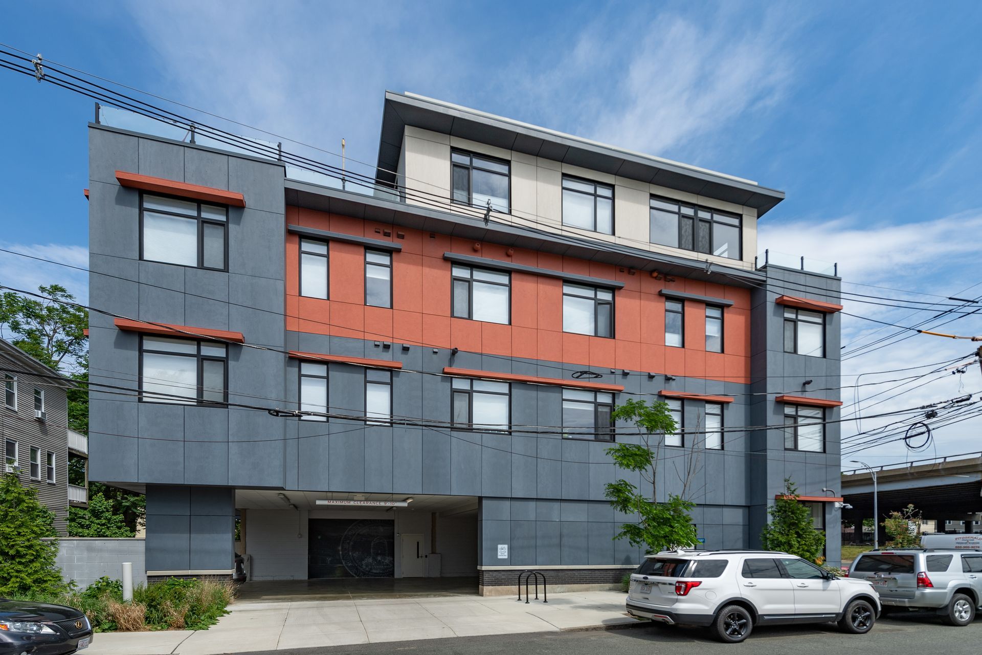 Modern gray and orange building with parking garage, cars parked in front, blue sky.