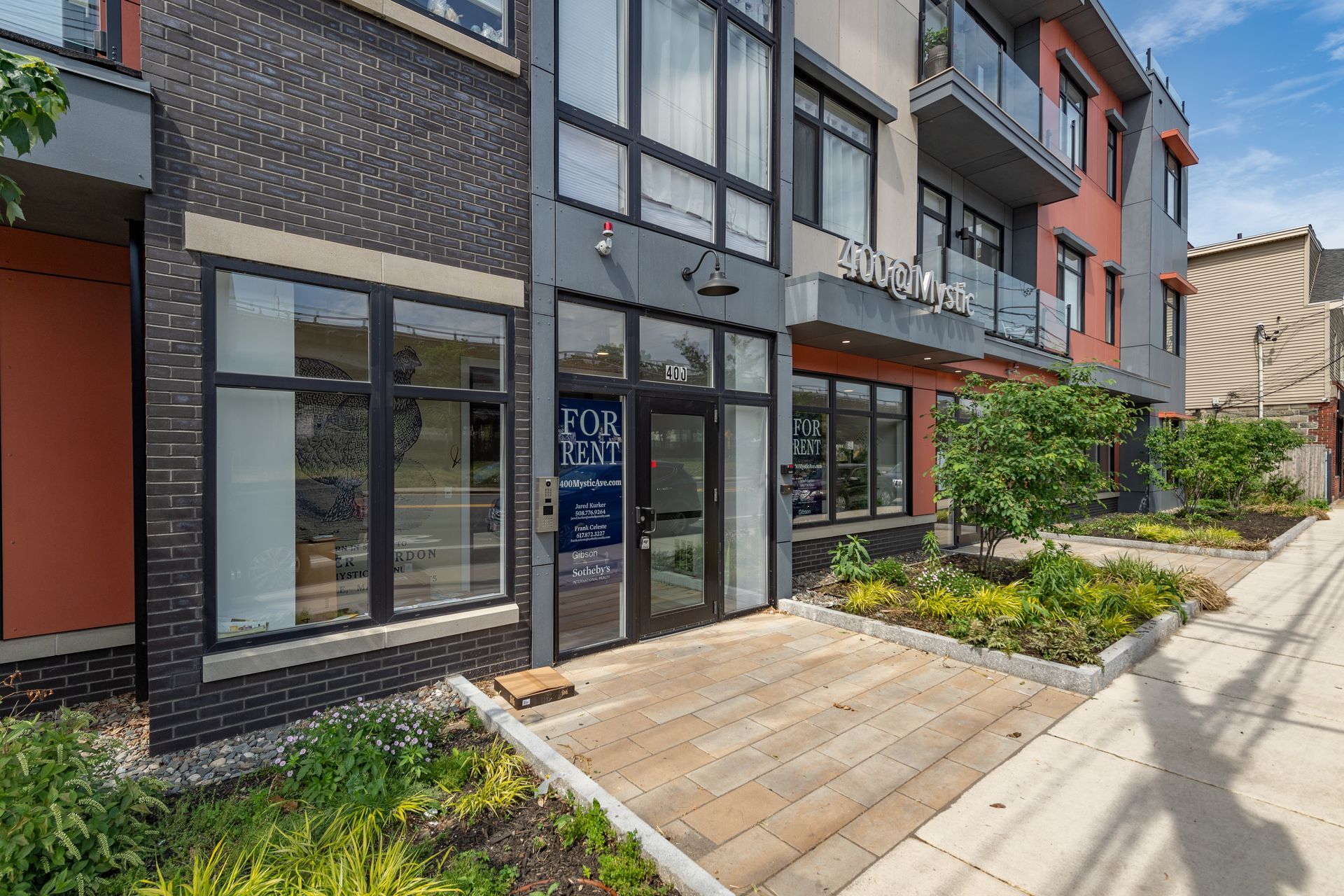 Exterior of a modern building with a storefront entrance and landscaping; gray, black, and orange color scheme.