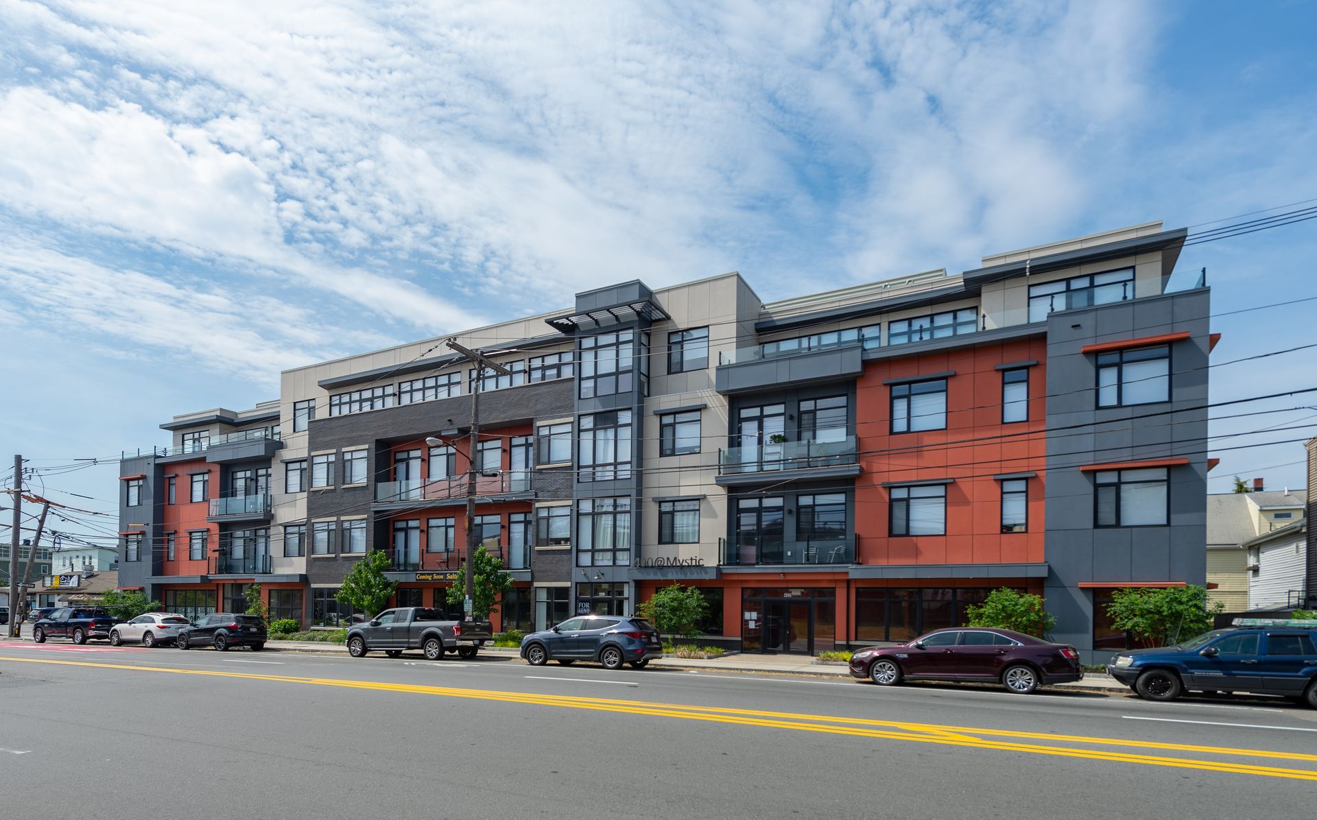 Modern multi-story apartment building with balconies, gray and orange facade, parked cars on street.