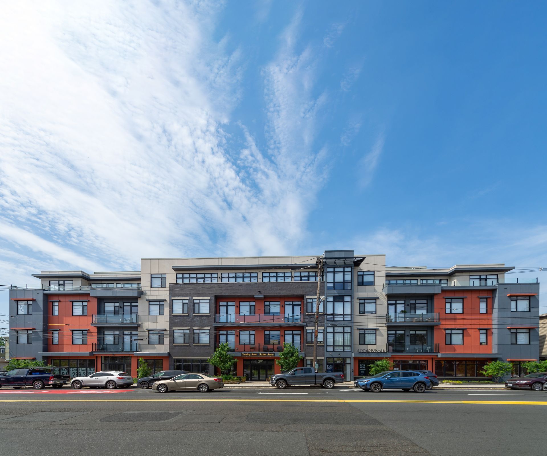 Multi-story building with balconies, varying brick colors, parked cars in front, and a blue sky with clouds.