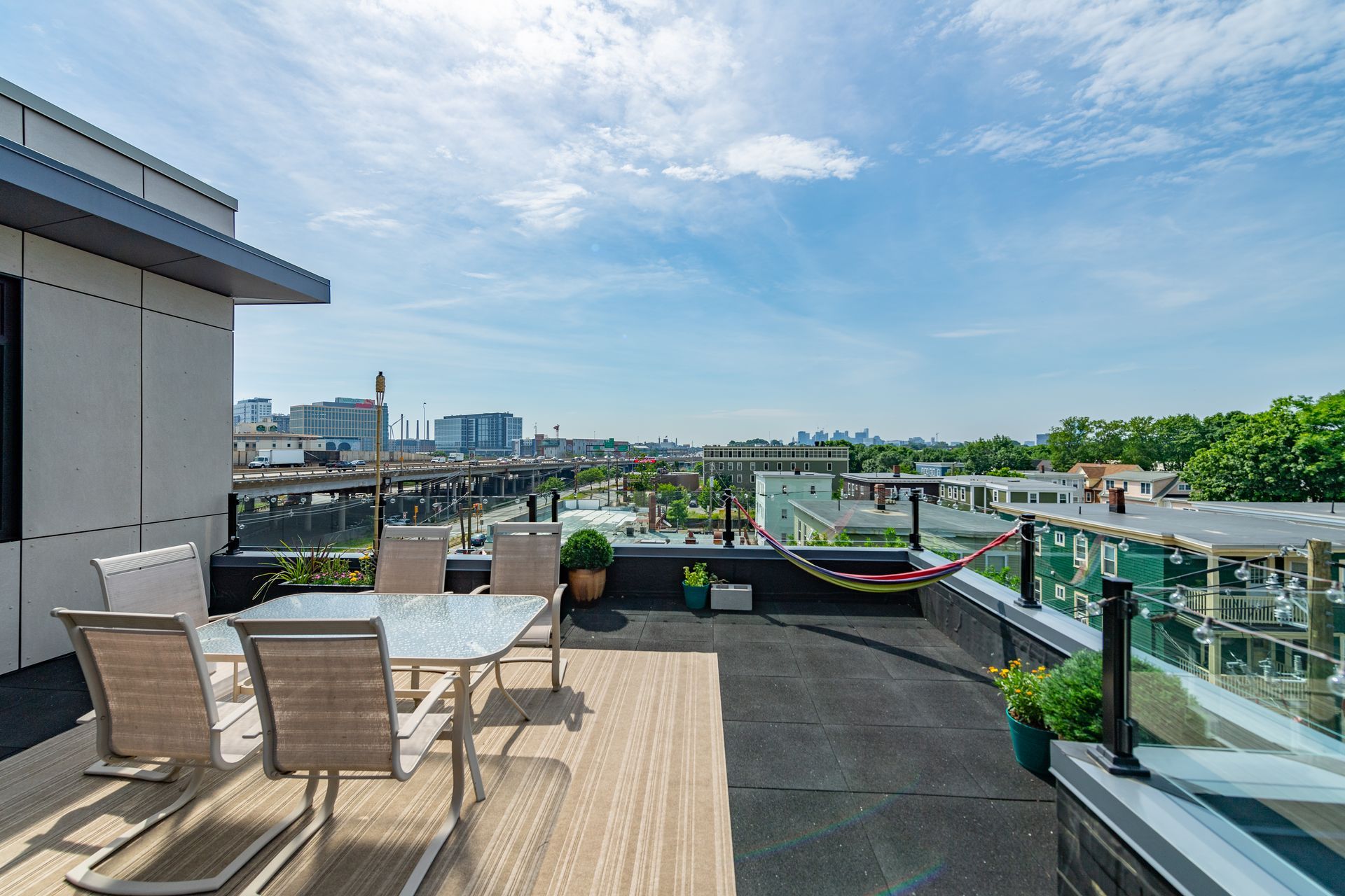 Rooftop patio with table, chairs, plants, and city view under a blue sky.