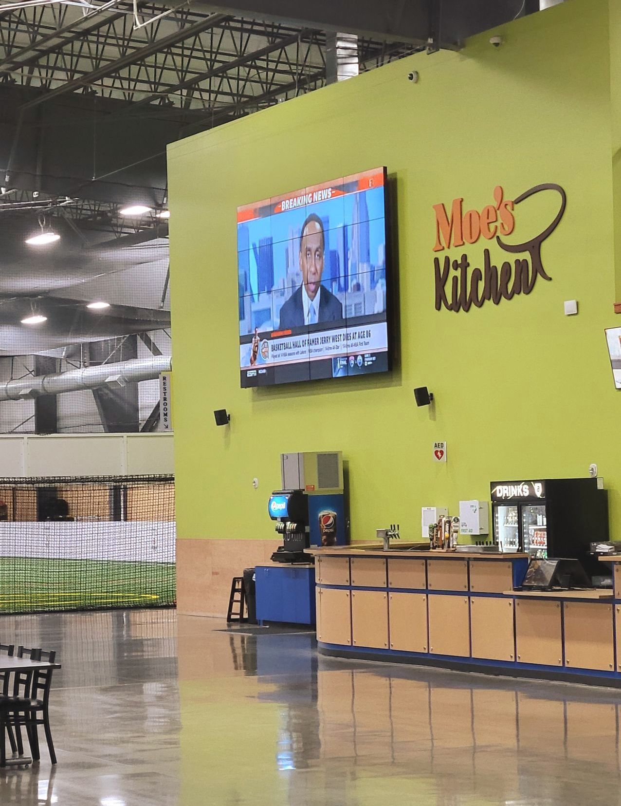 Interior of Moe's Kitchen with a TV showing a news anchor. Green wall with counter and refrigerators.