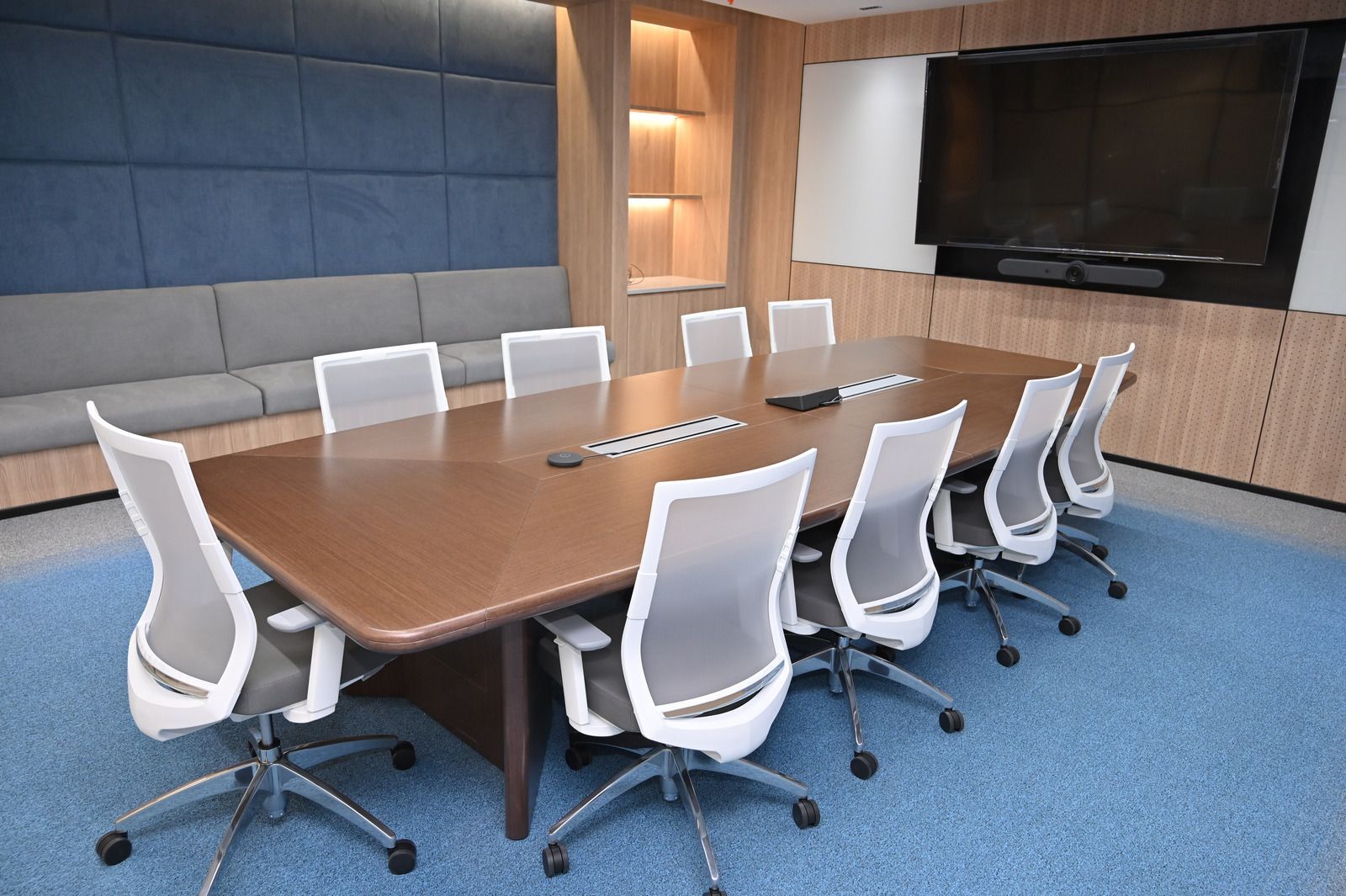 A modern conference room with a large wooden table, grey mesh chairs, a blue upholstered wall, and a mounted screen.