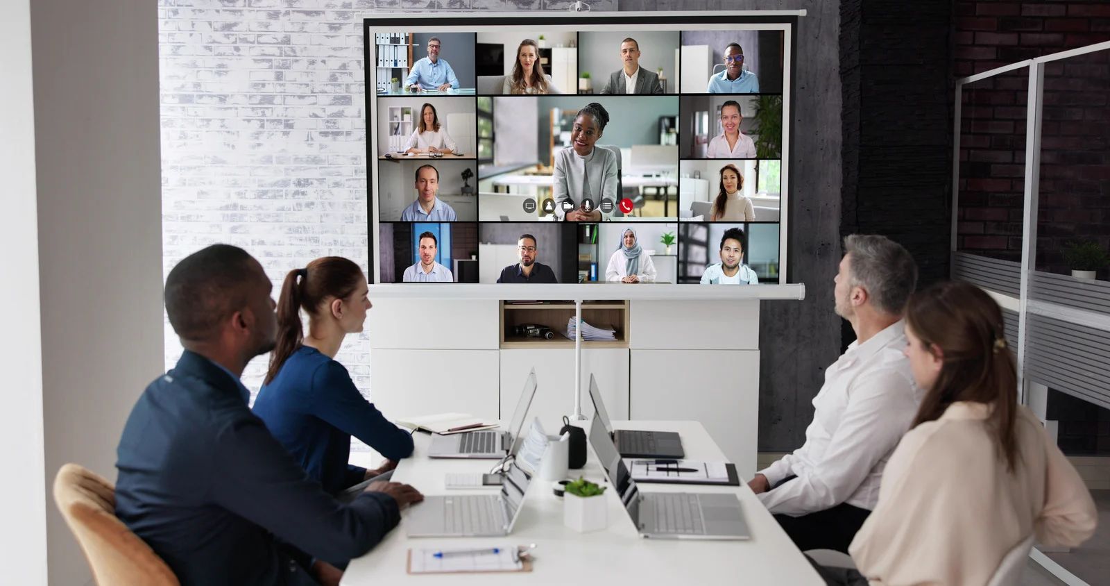 Four people sit at a table facing a large monitor displaying a video conference with many participants in an office.
