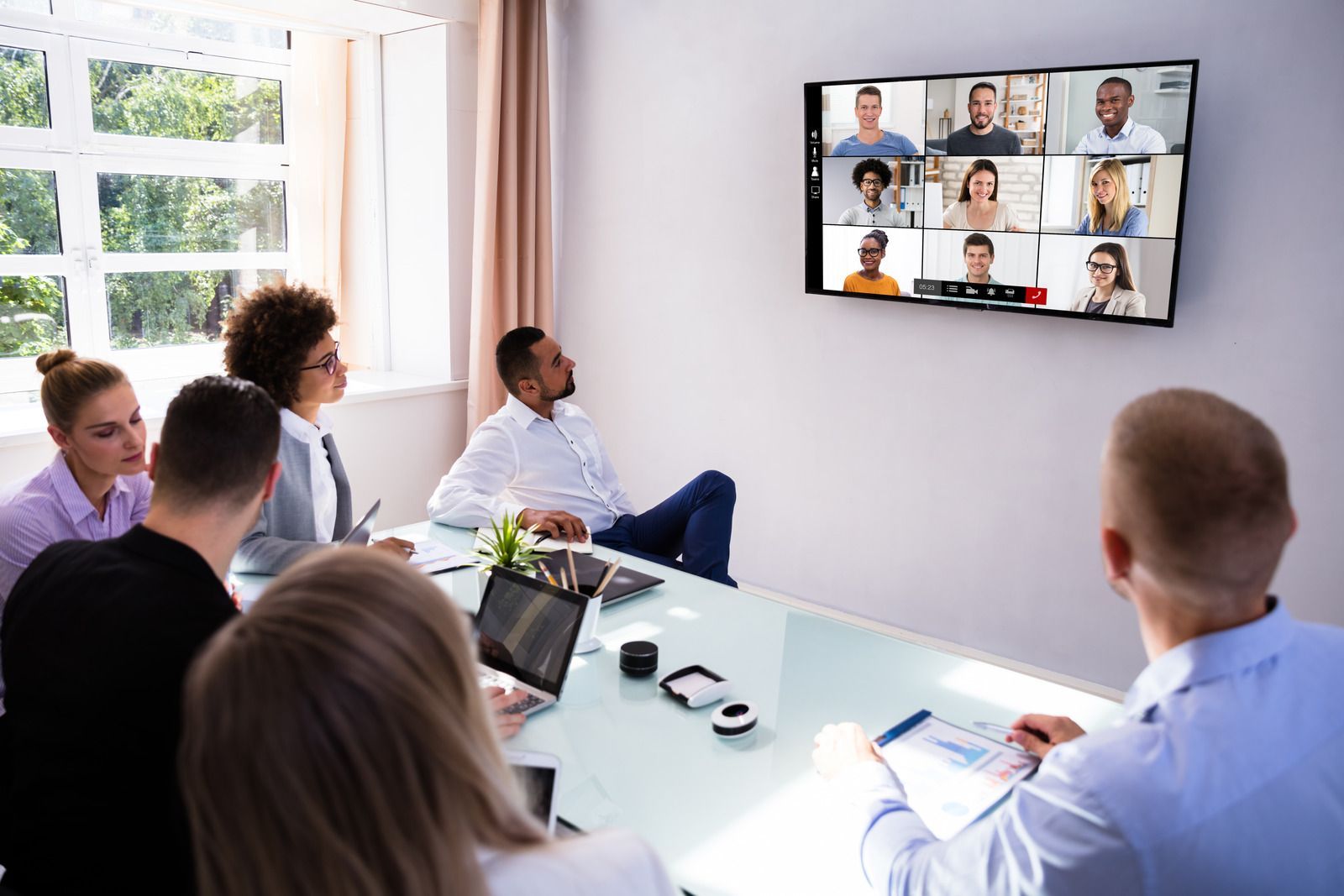 People in a conference room watching a video call on a wall-mounted screen, a window on the left.