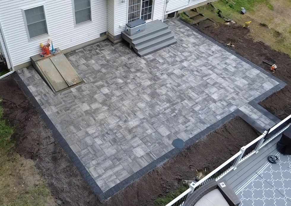 Brick patio adjacent to a white house with steps. Dark border, some dirt, and partially visible deck.