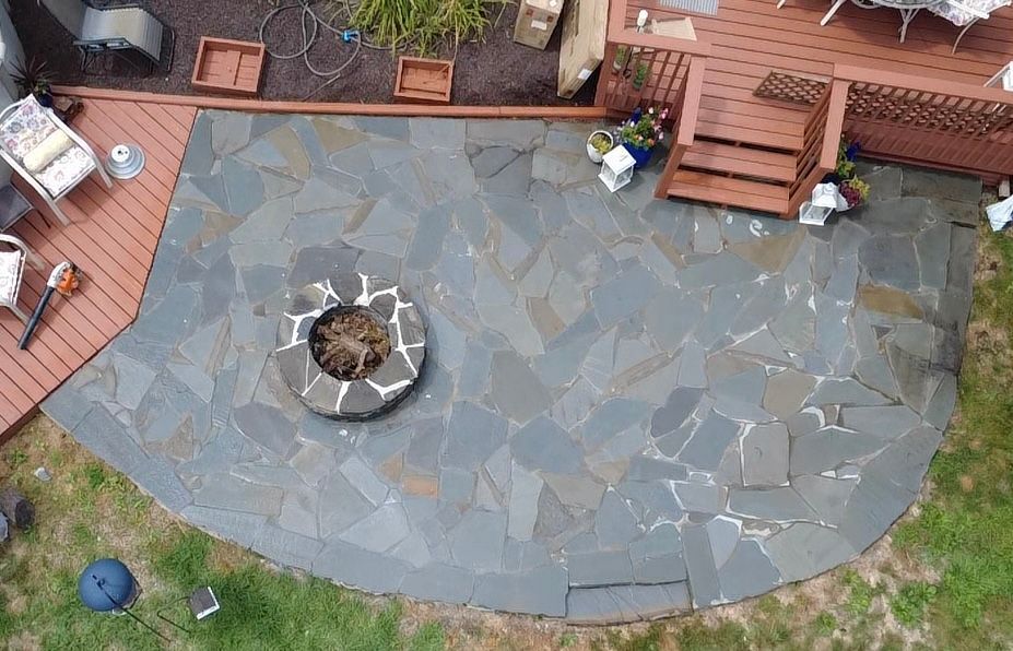 Overhead view of a stone patio with a fire pit, adjacent to a wooden deck and bordered by grass.