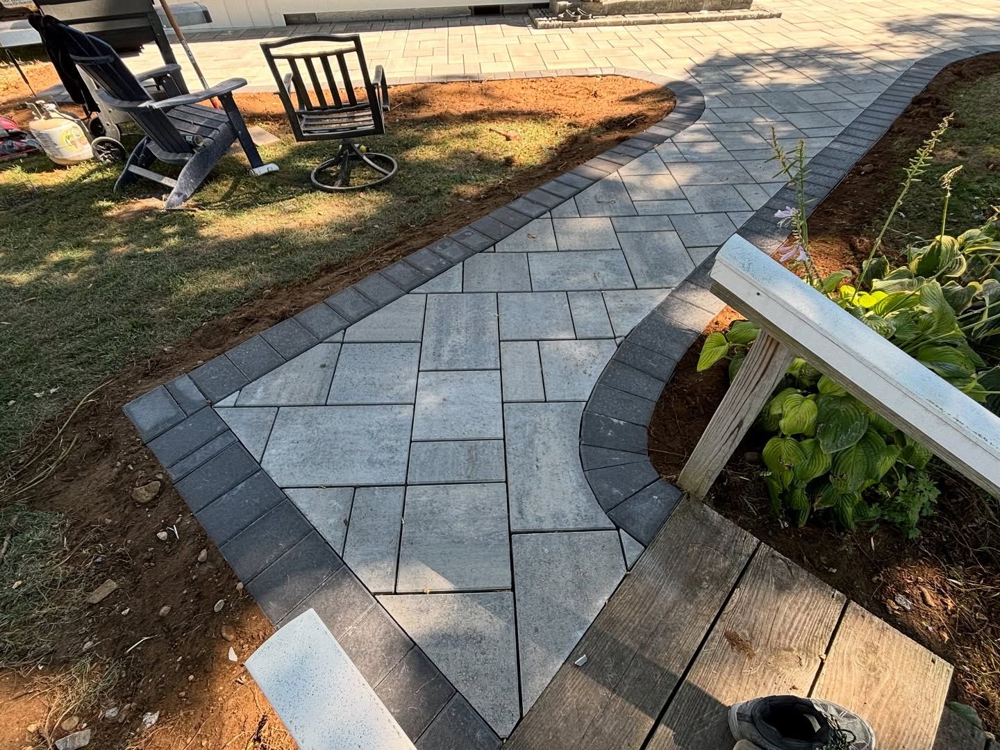 Curved stone garden path with brick edging beside lawn, plants, and outdoor chairs in sunlight
