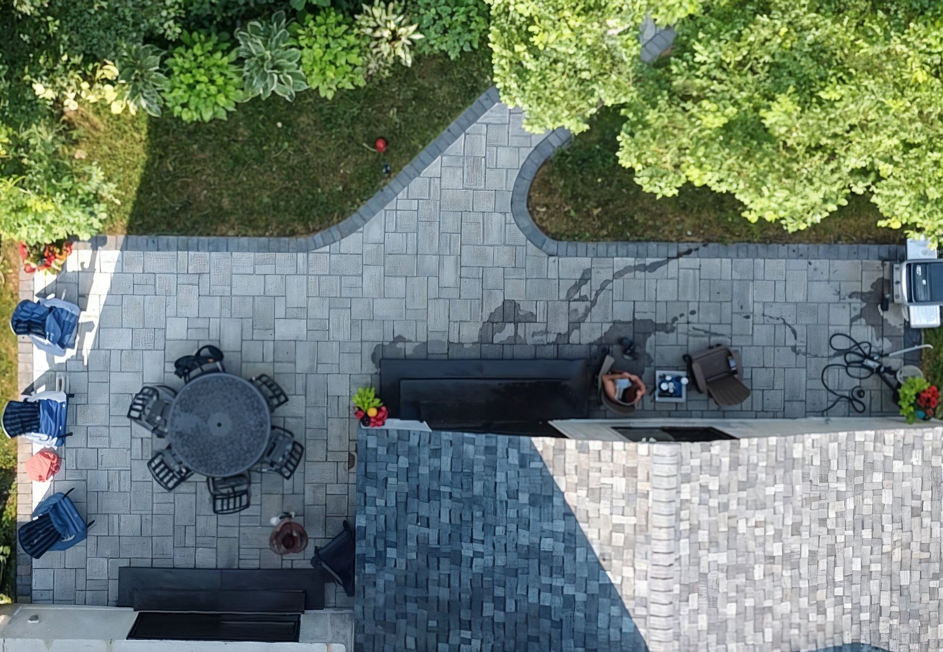 Overhead view of a backyard patio with a stone surface, furniture, and greenery. A car is parked on the right.