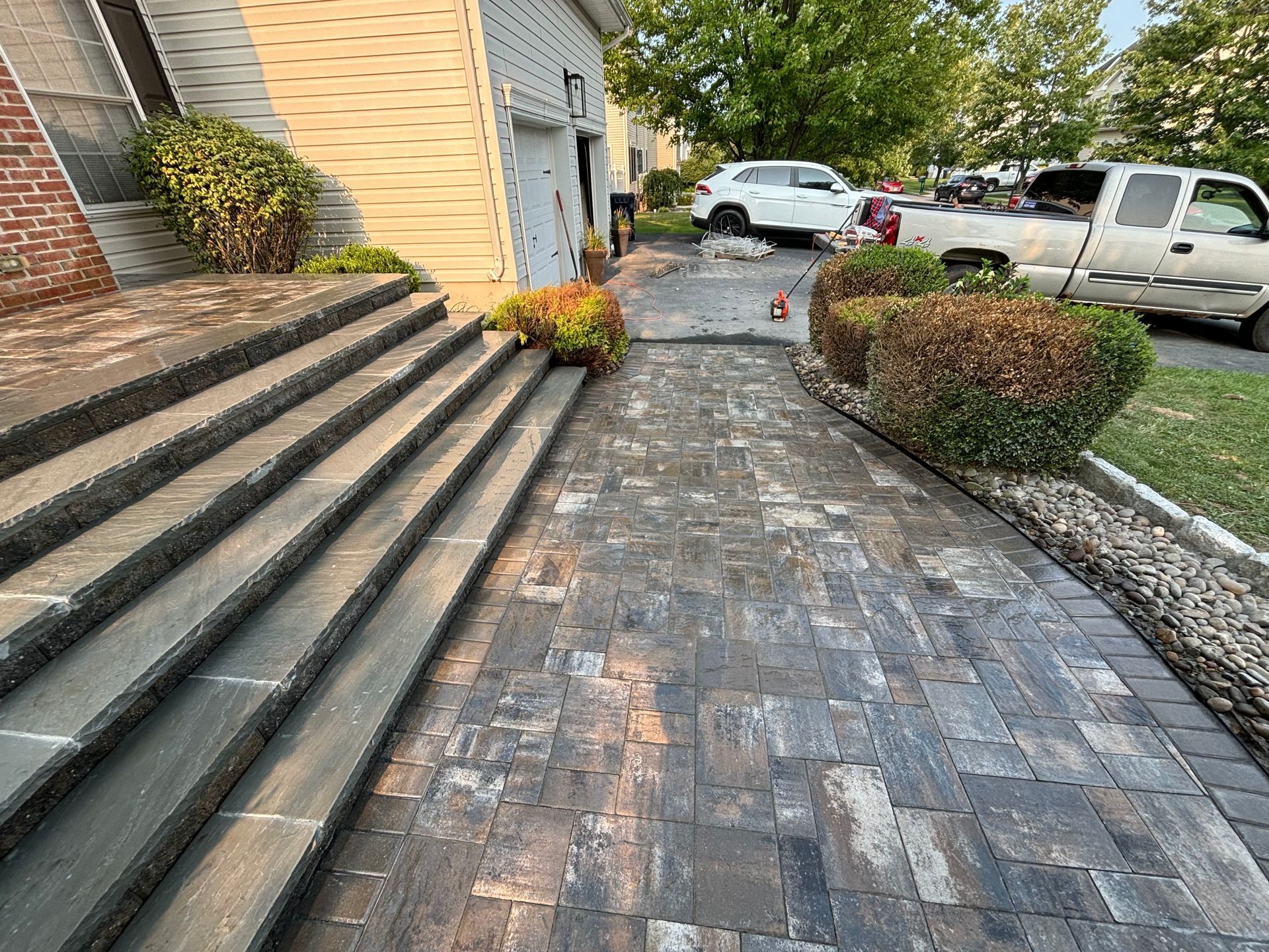 Brick pathway and steps leading to a house, flanked by bushes and a driveway with vehicles.