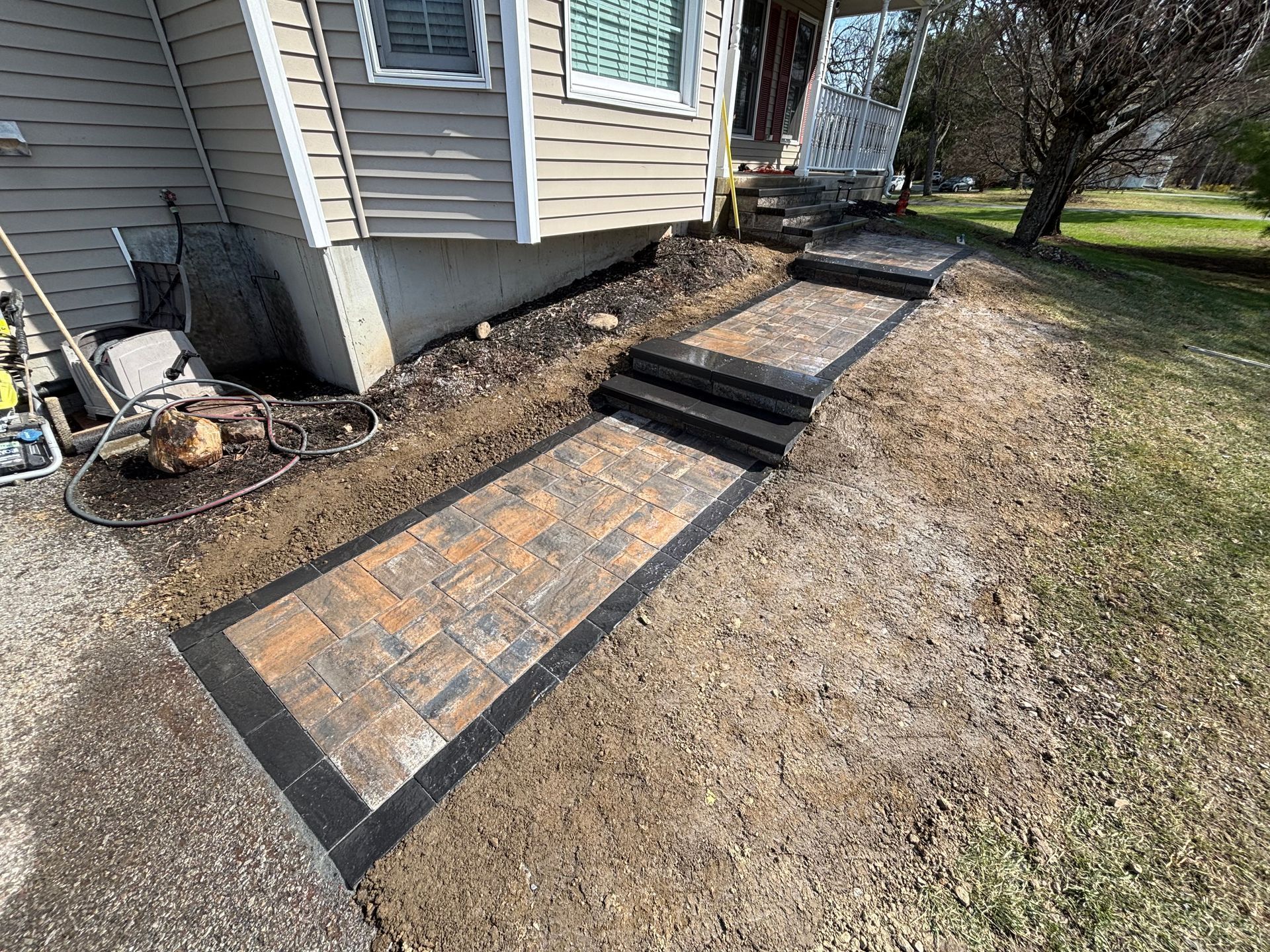 Brick pathway with steps leading up to a house entrance; black border and tan siding on the house.