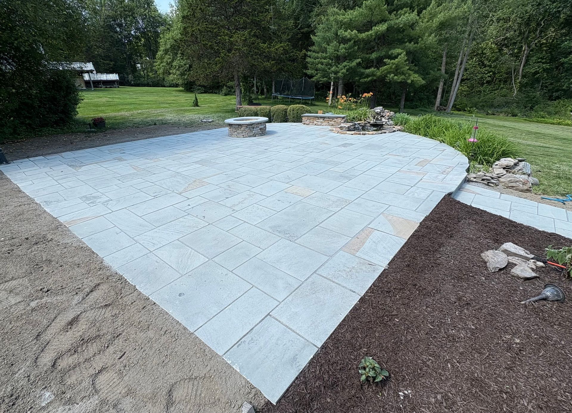 Light gray paved patio in a yard with a fire pit, surrounded by mulch and grass.