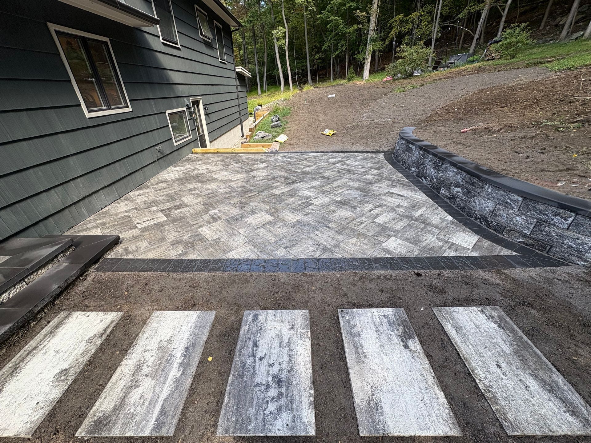 Stone patio with stepping stones leading to a house, surrounded by a retaining wall and wooded area.