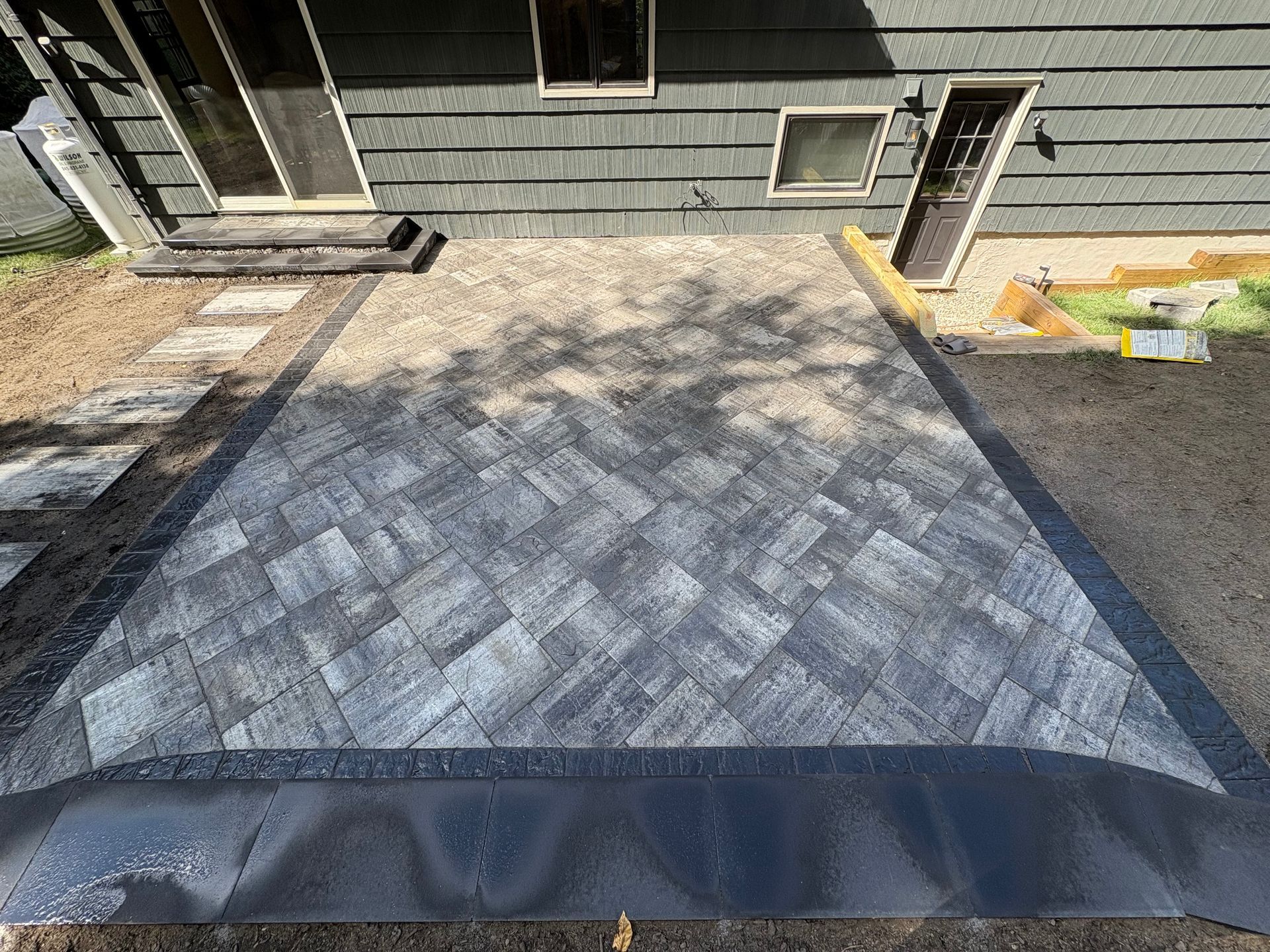 Stone patio with herringbone pattern, black border, next to a gray house with doors and windows.