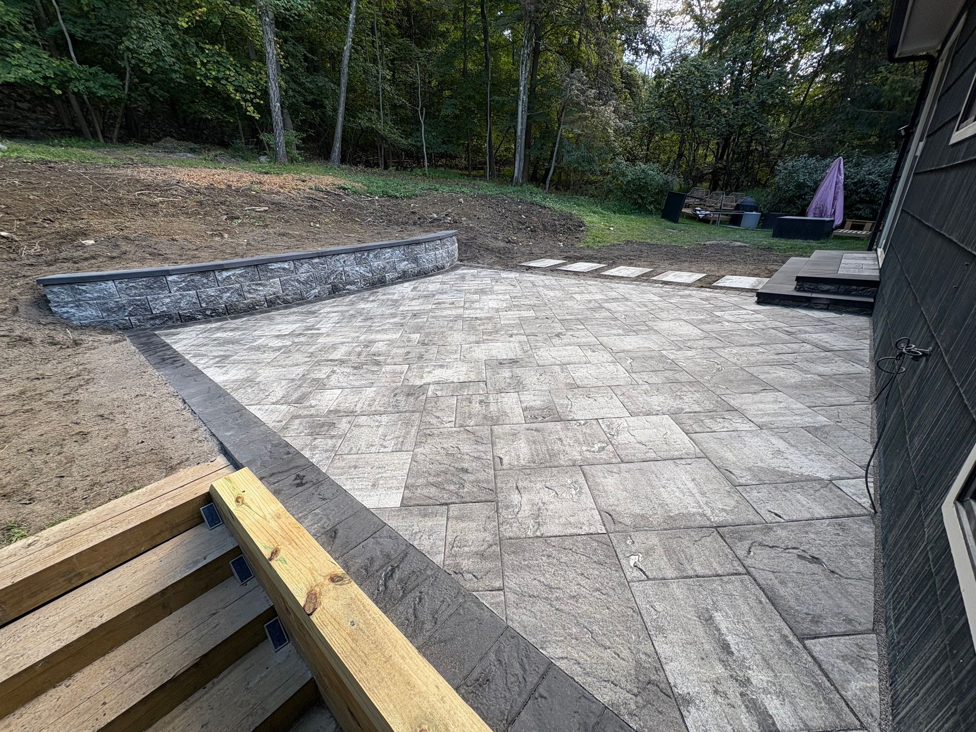 Patio with gray pavers, retaining wall, and wooden structure; set in a yard with trees.