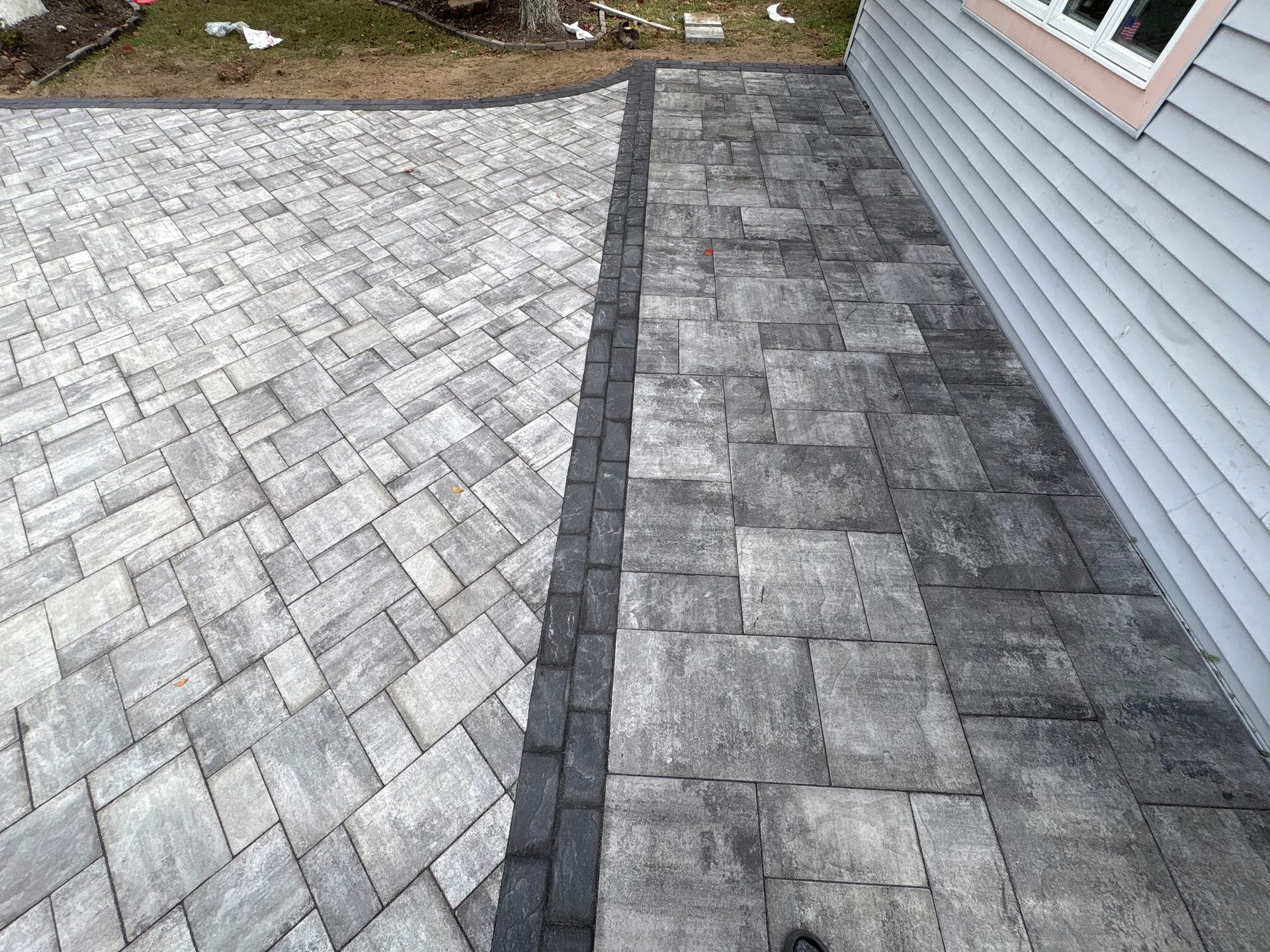 Gray and dark gray paved patio with a black border, next to a house with gray siding.