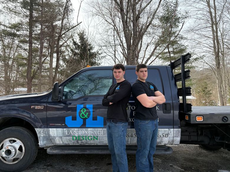 Two men with arms crossed stand by a blue pickup truck with 