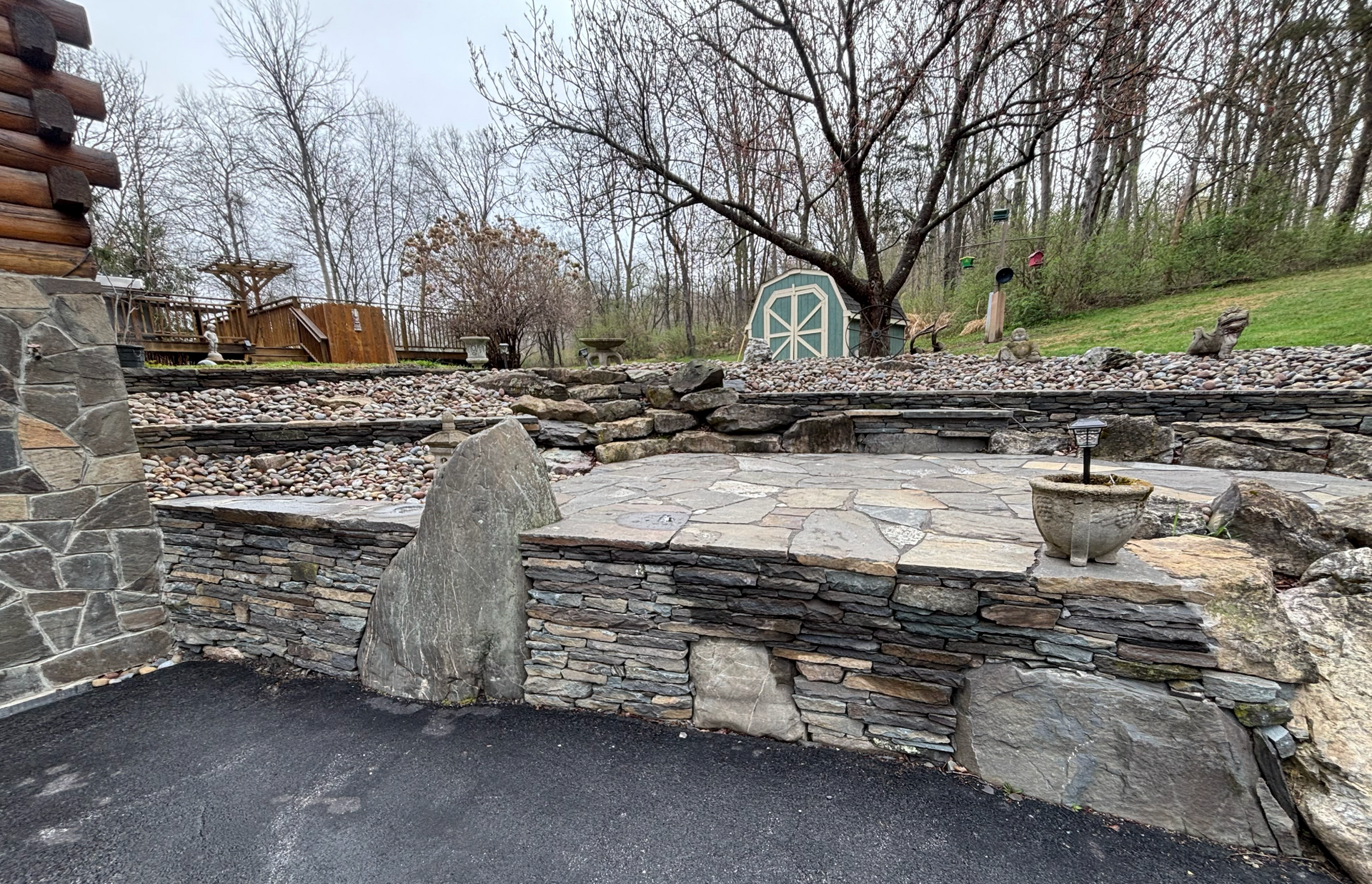 Stone patio with tiered levels, gravel beds, and trees in background. Cloudy sky.