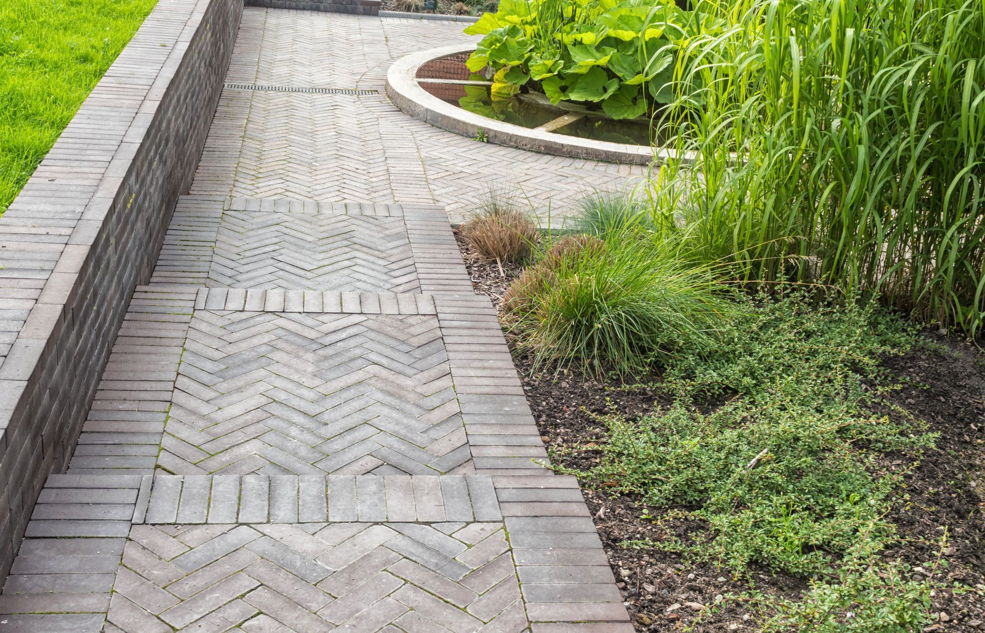 Curved brick walkway beside a landscaped garden with tall green plants and grass