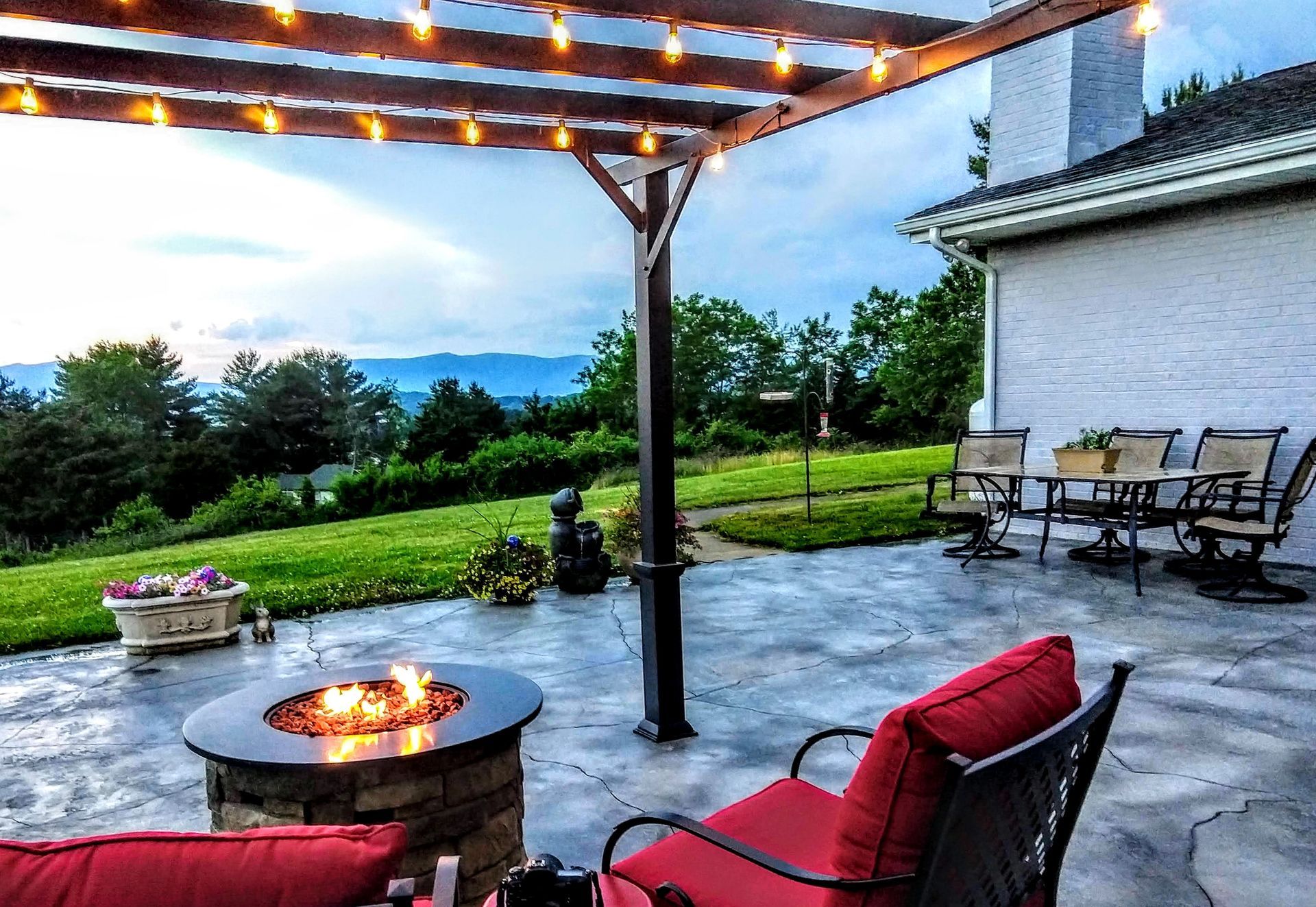 Covered patio with string lights, fire pit, red chairs, and a grassy yard at sunset