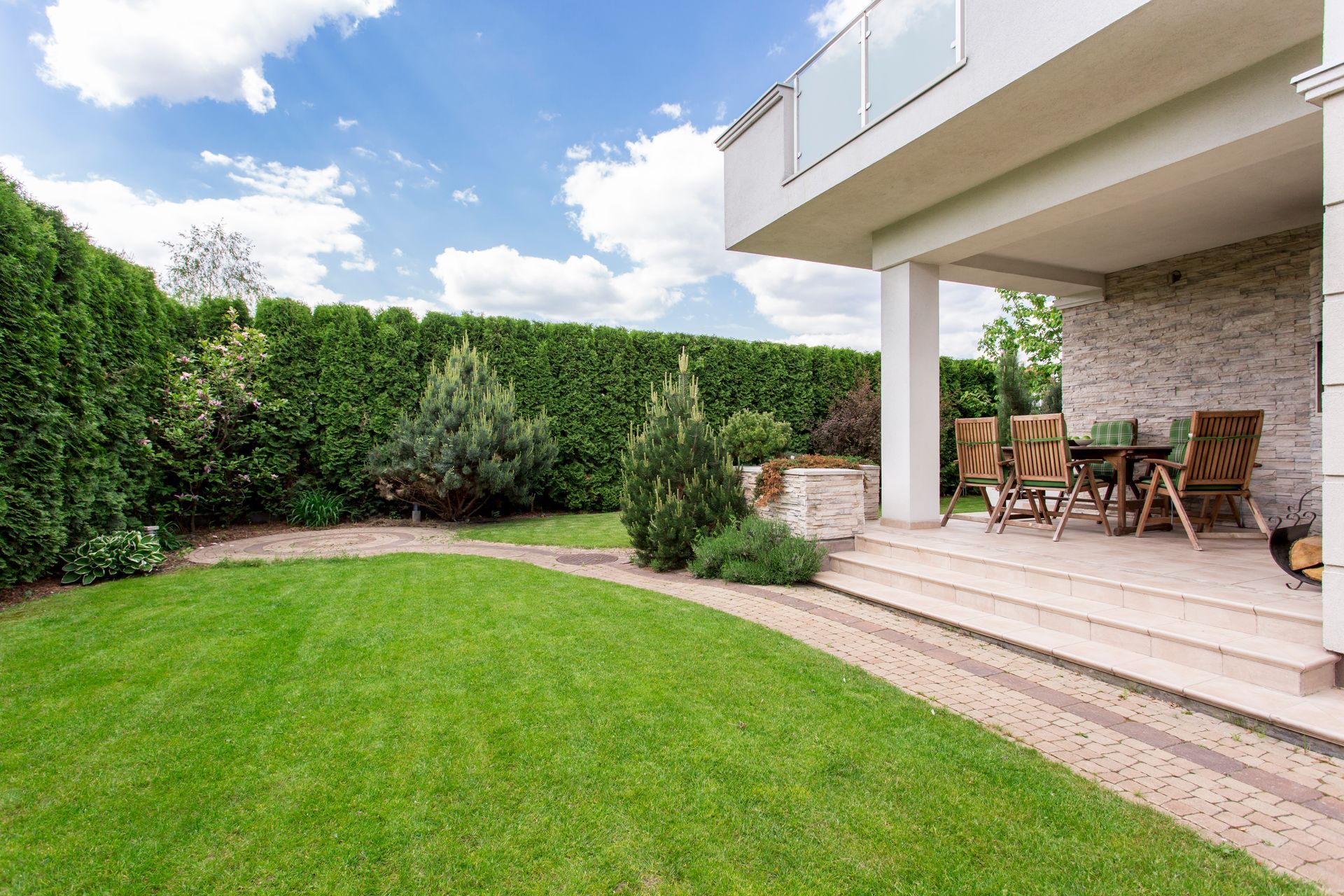 Modern backyard patio with wooden lounge chairs, stone steps, and a green lawn beside a hedge