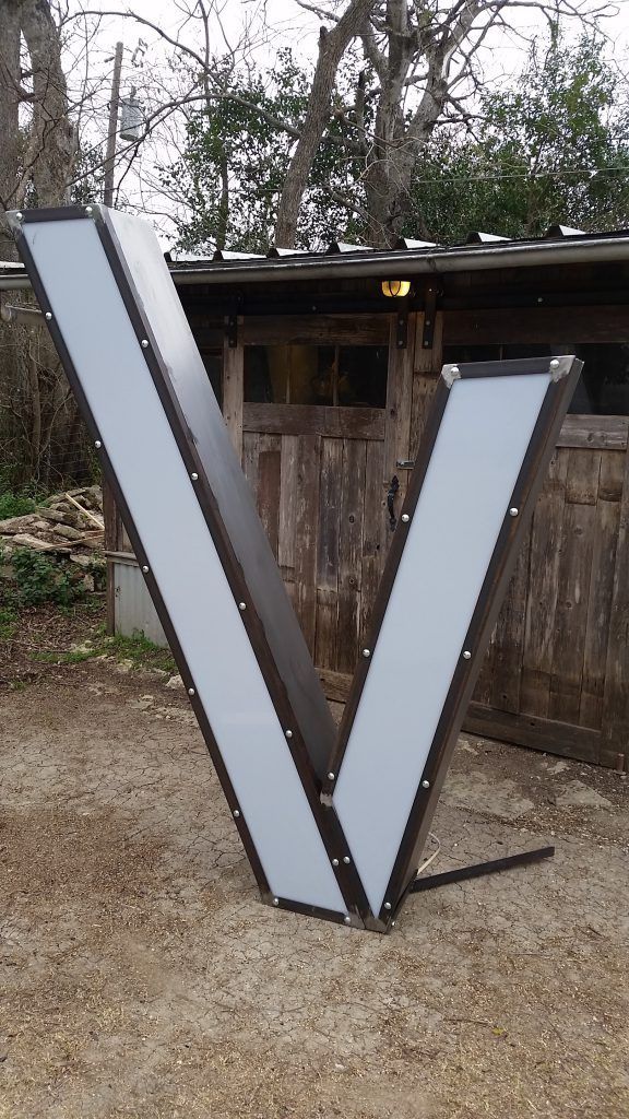 A large letter v is sitting in the dirt in front of a wooden shed.