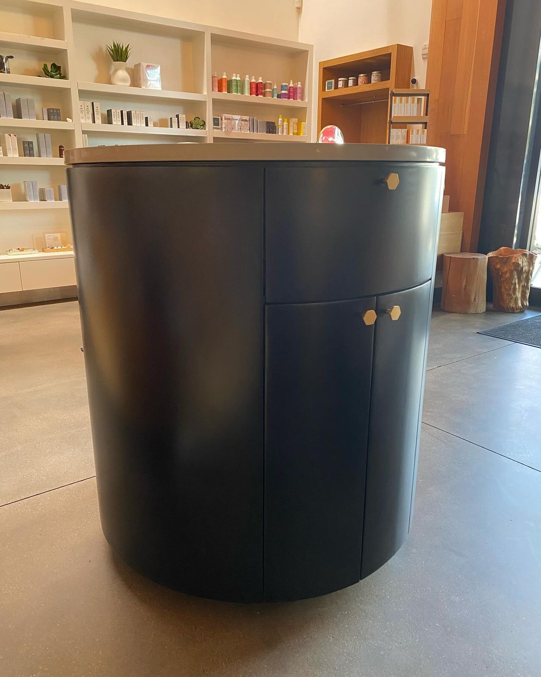A round black cabinet with gold handles is in a room with shelves.