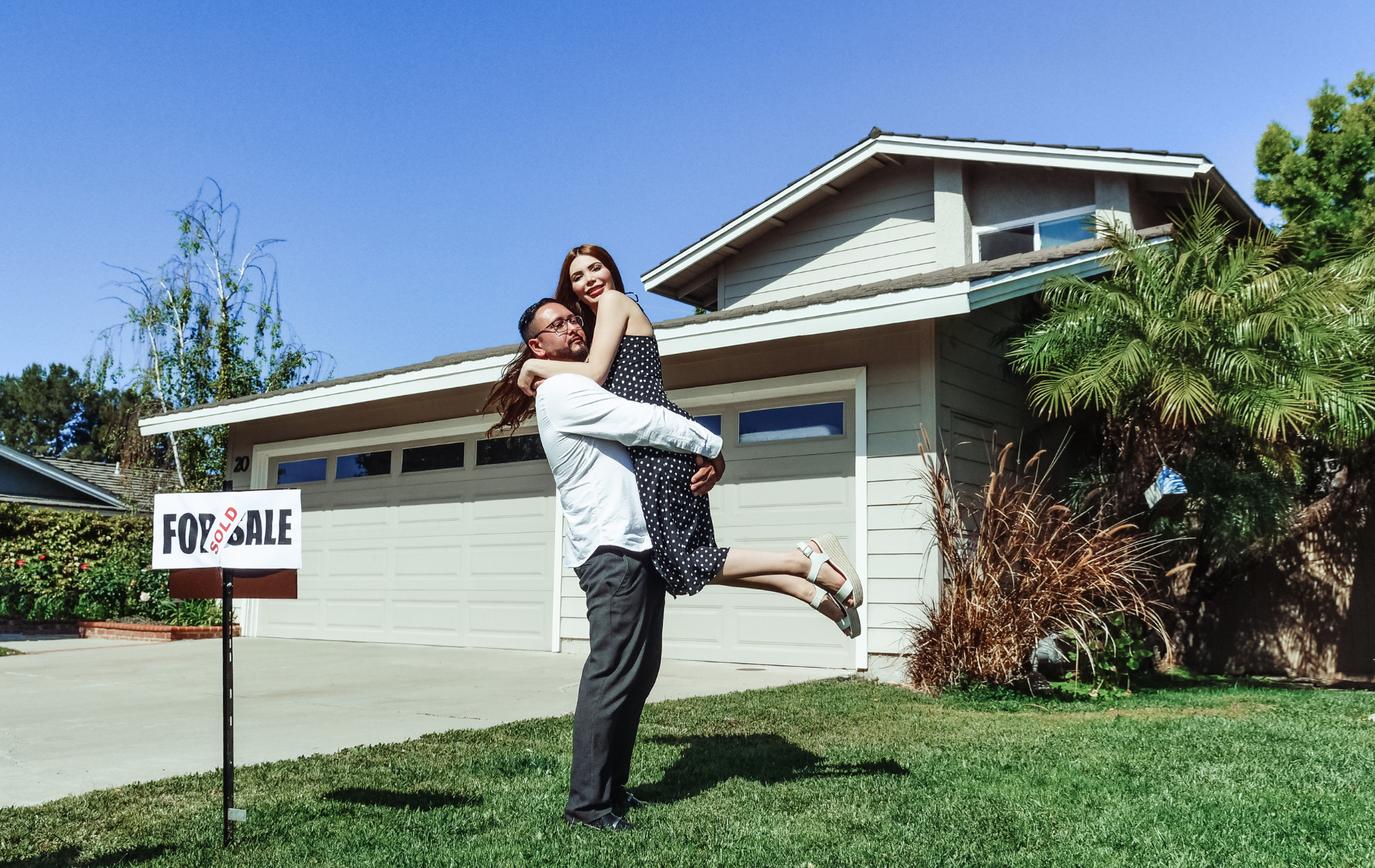A man is holding a woman in his arms in front of a for sale sign.