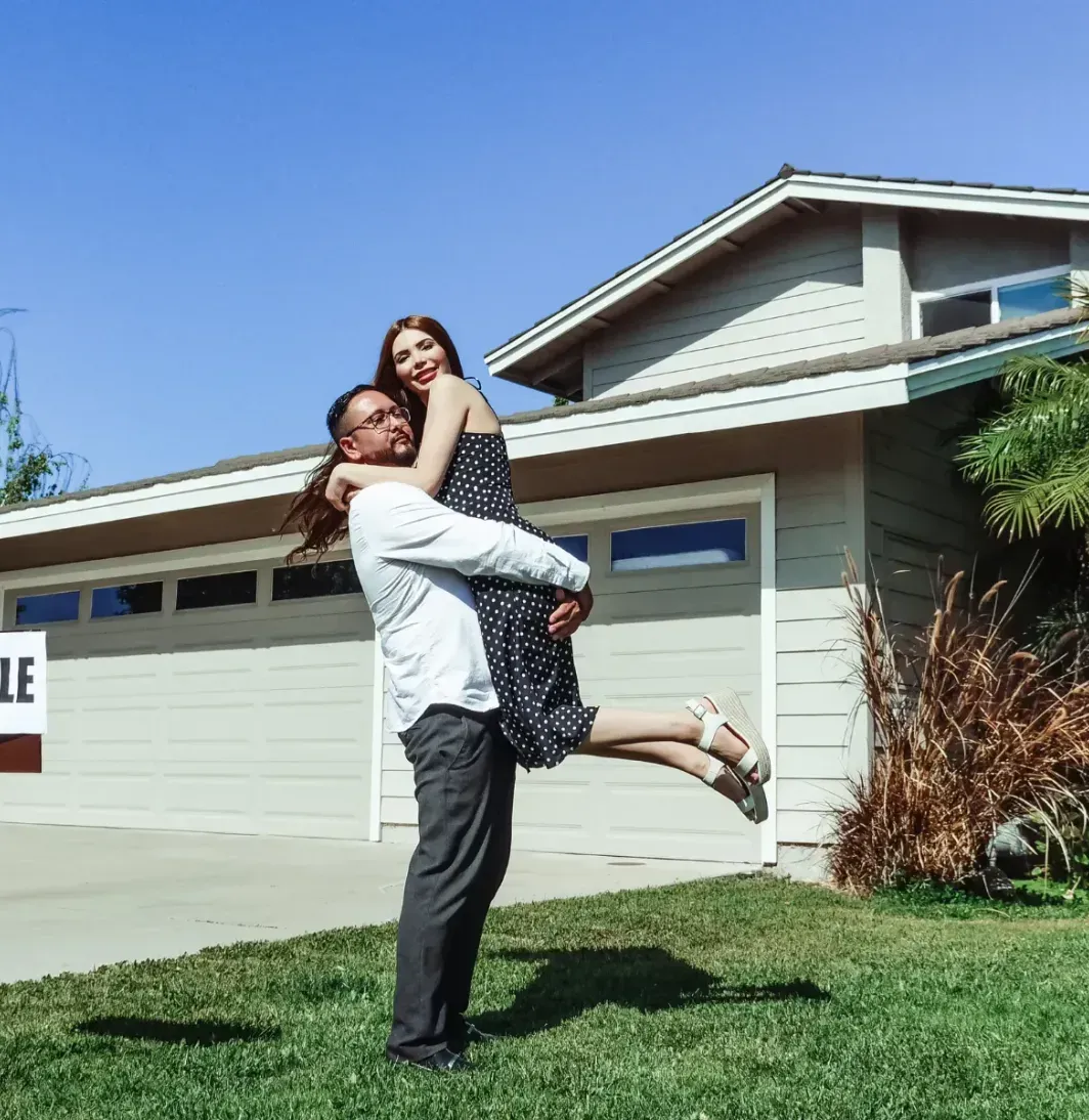 A man is holding a woman in his arms in front of a for sale sign.