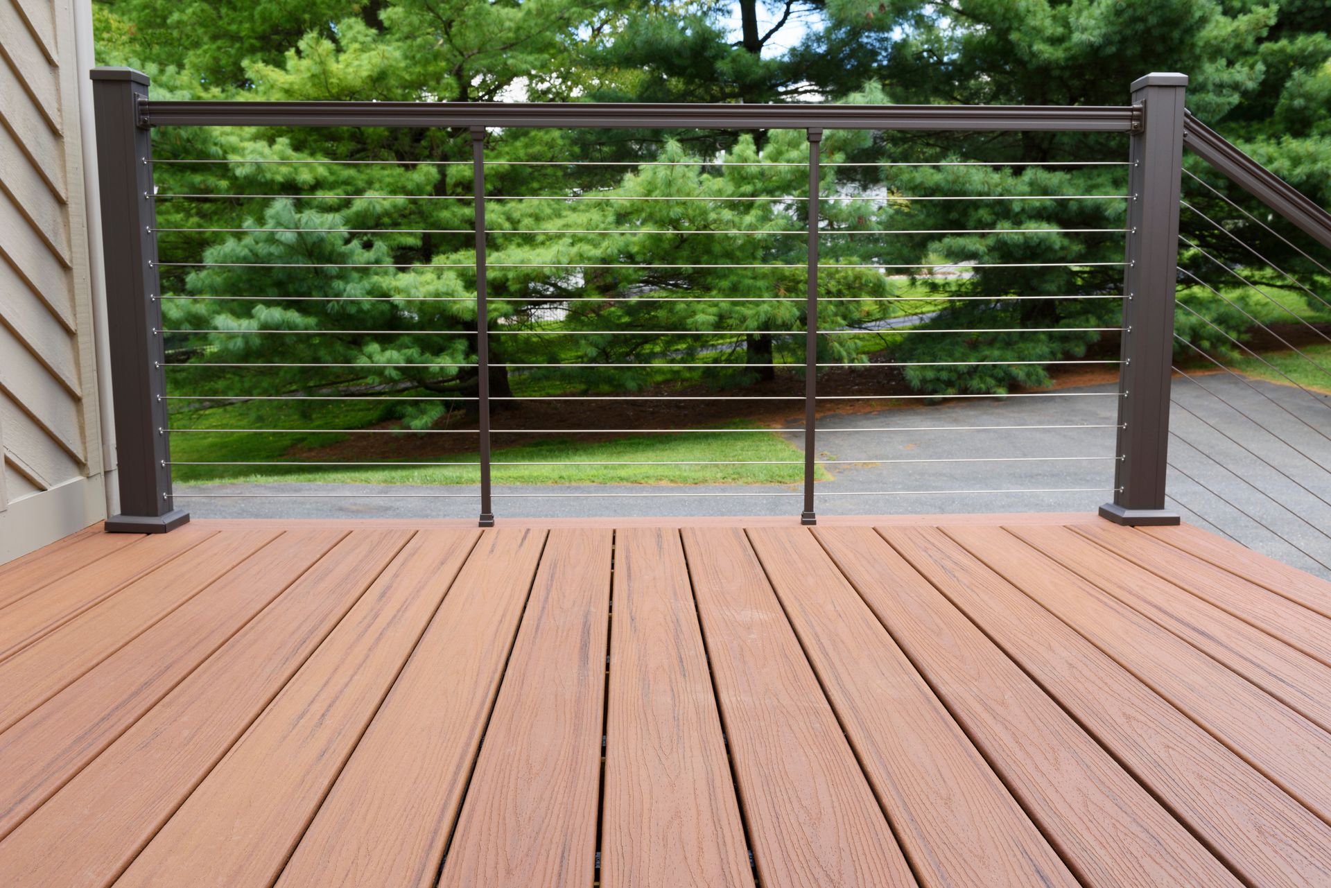 Wooden deck with horizontal cable railing overlooking a green backyard with trees and a paved area.