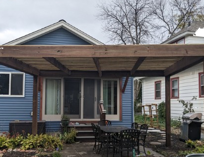 Pergola over a patio with a table and chairs, adjacent to a blue house with sliding doors.