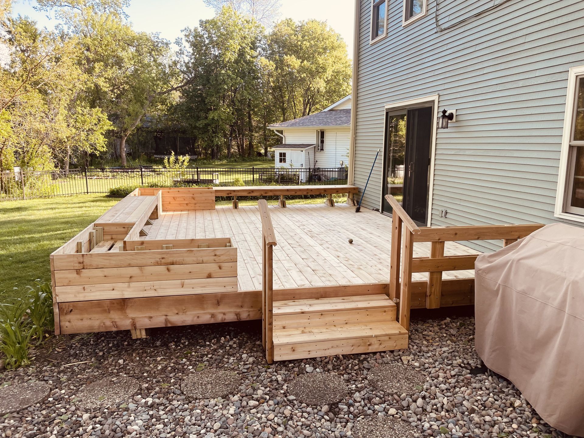 Wooden deck with built-in seating, stairs, and a ramp, next to a light blue house. Overlooking a yard with trees.