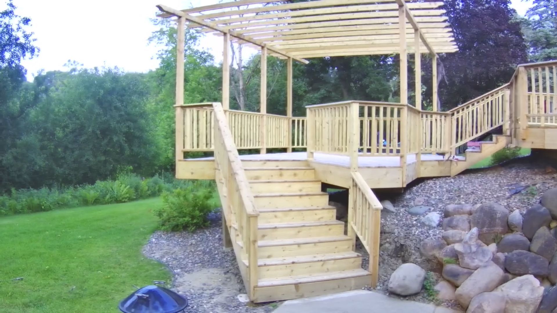 Wooden deck with stairs, pergola, and railing in a yard with grass and rock landscaping.