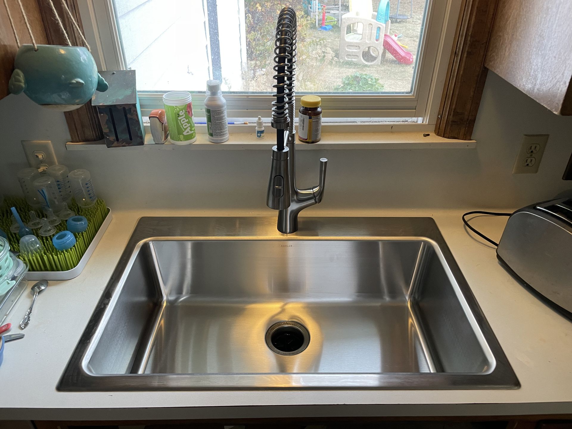Stainless steel kitchen sink with a tall faucet, beside a countertop with a window and toaster.